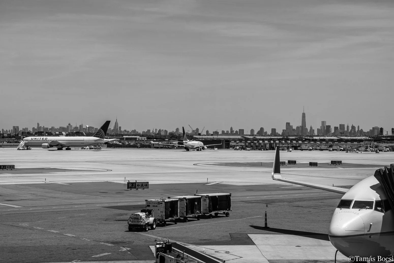 Black and white photo of an airport tarmac with airplane parts and baggage carts, with a city skyline featuring the Empire State Building in the background.