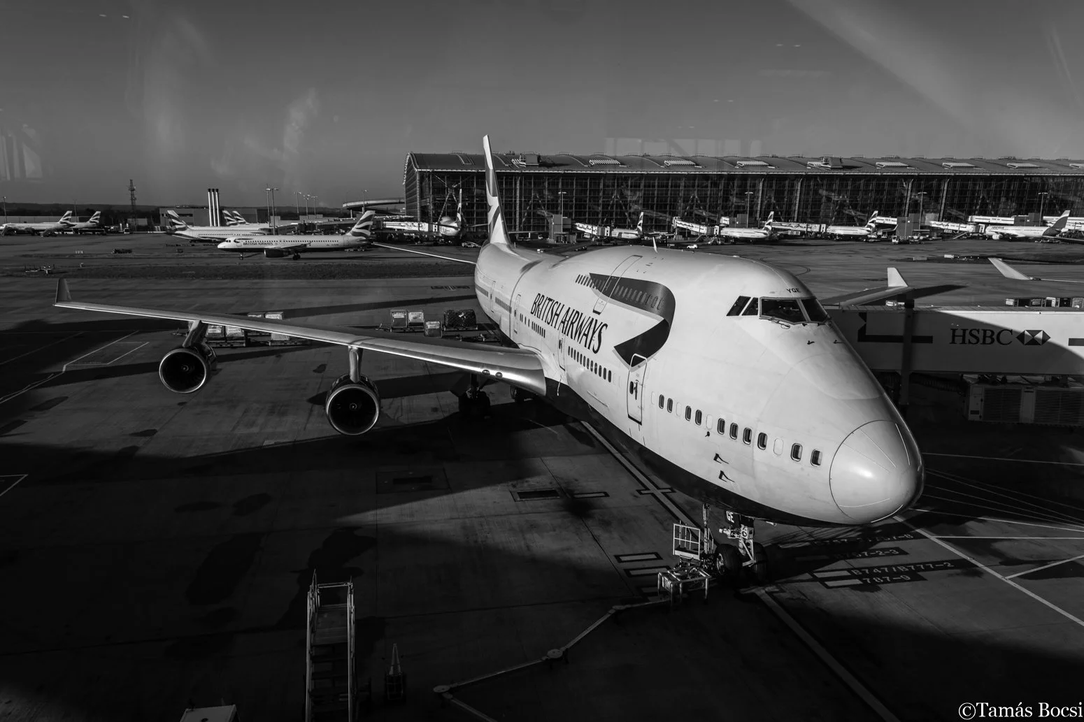 A British Airways airplane at an airport gate, with other aircraft and airport structures visible in the background.