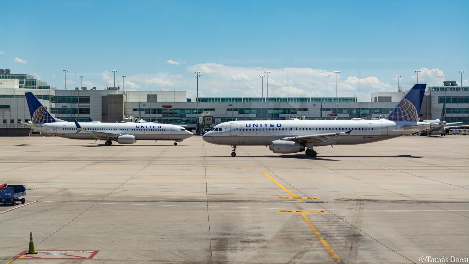 Two United Airlines airplanes parked on an airport tarmac in front of a terminal building with glass windows, under a partly cloudy sky.
