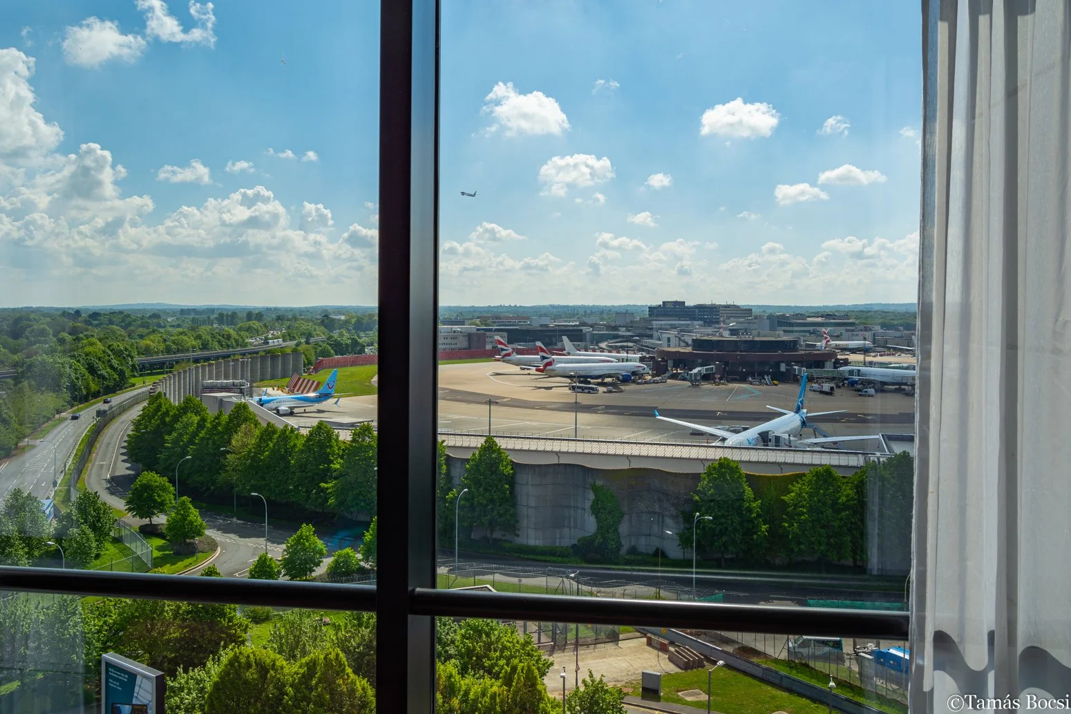 View of an airport runway and several airplanes parked at the gates, seen through a large window with white curtains, with a blue sky and scattered clouds.