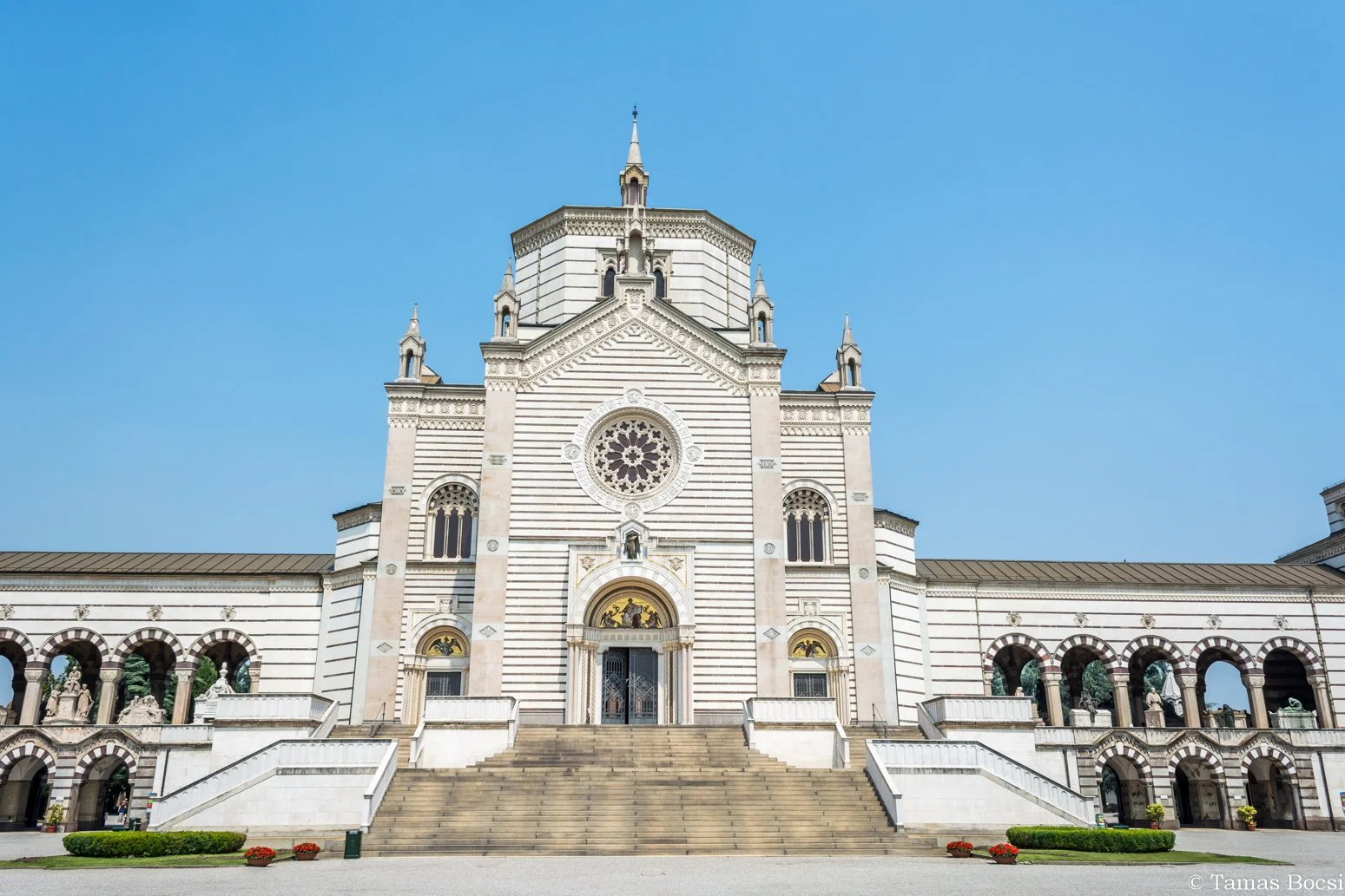 Front view of a large white and gray cathedral with stairs leading up to the main entrance, featuring a rose window and small towers on its facade, under a clear blue sky.