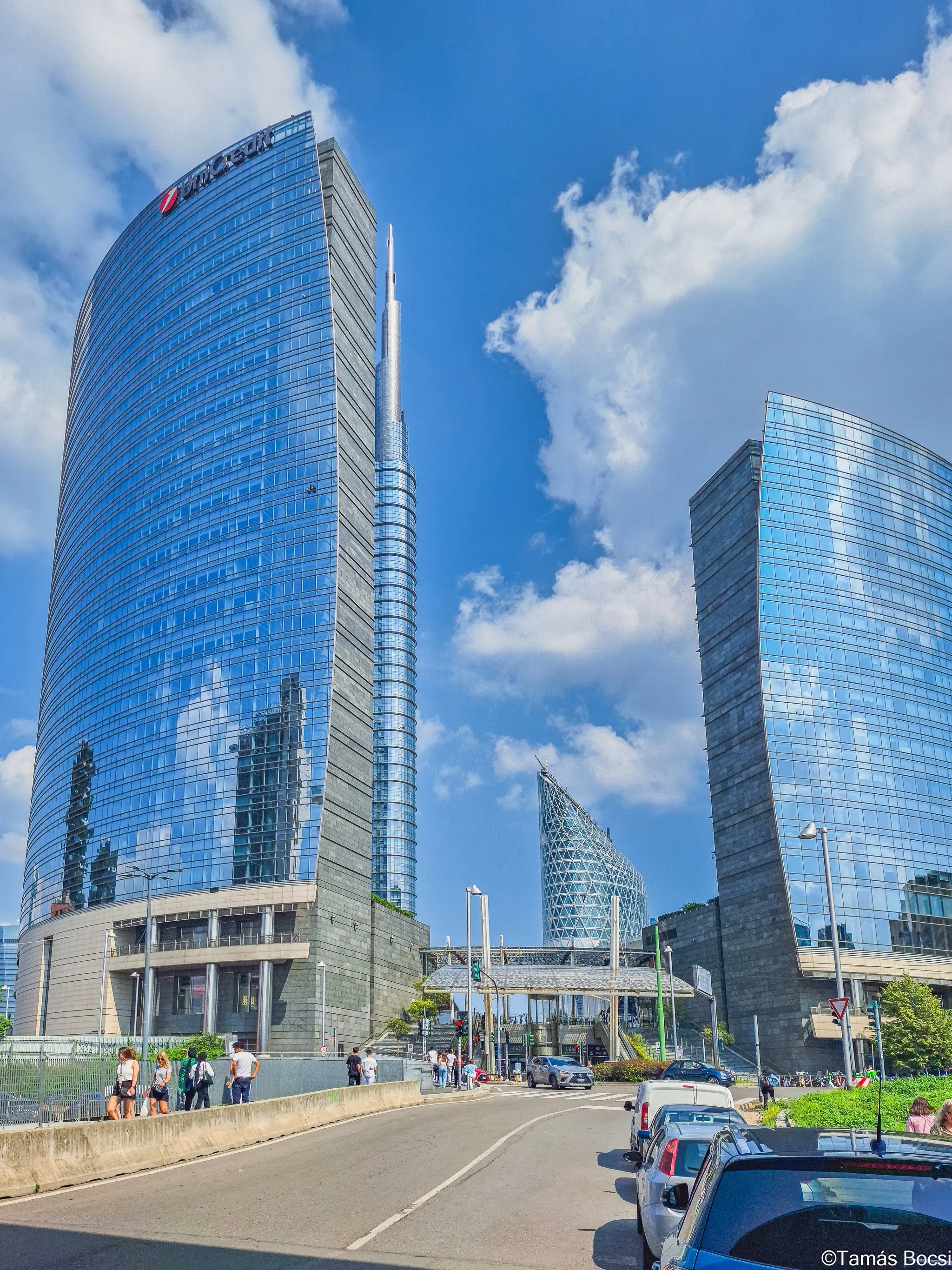 A cityscape with modern skyscrapers, including the tallest one with a pointed spire, under a partly cloudy blue sky, with people walking on a sidewalk and cars parked along the street.