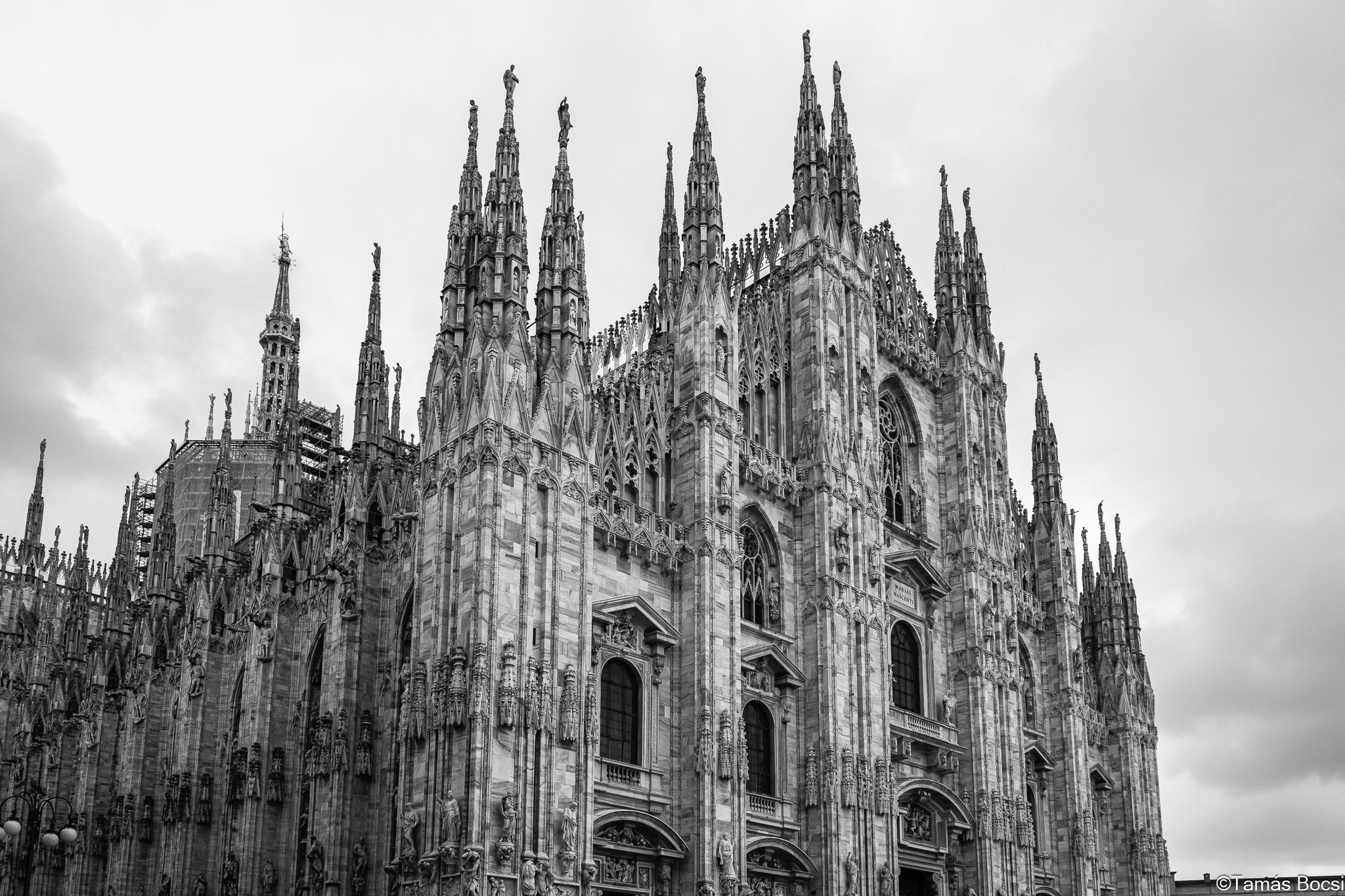 Black and white photo of the Milan Cathedral, an ornate Gothic church with multiple tall spires and detailed stone carvings.