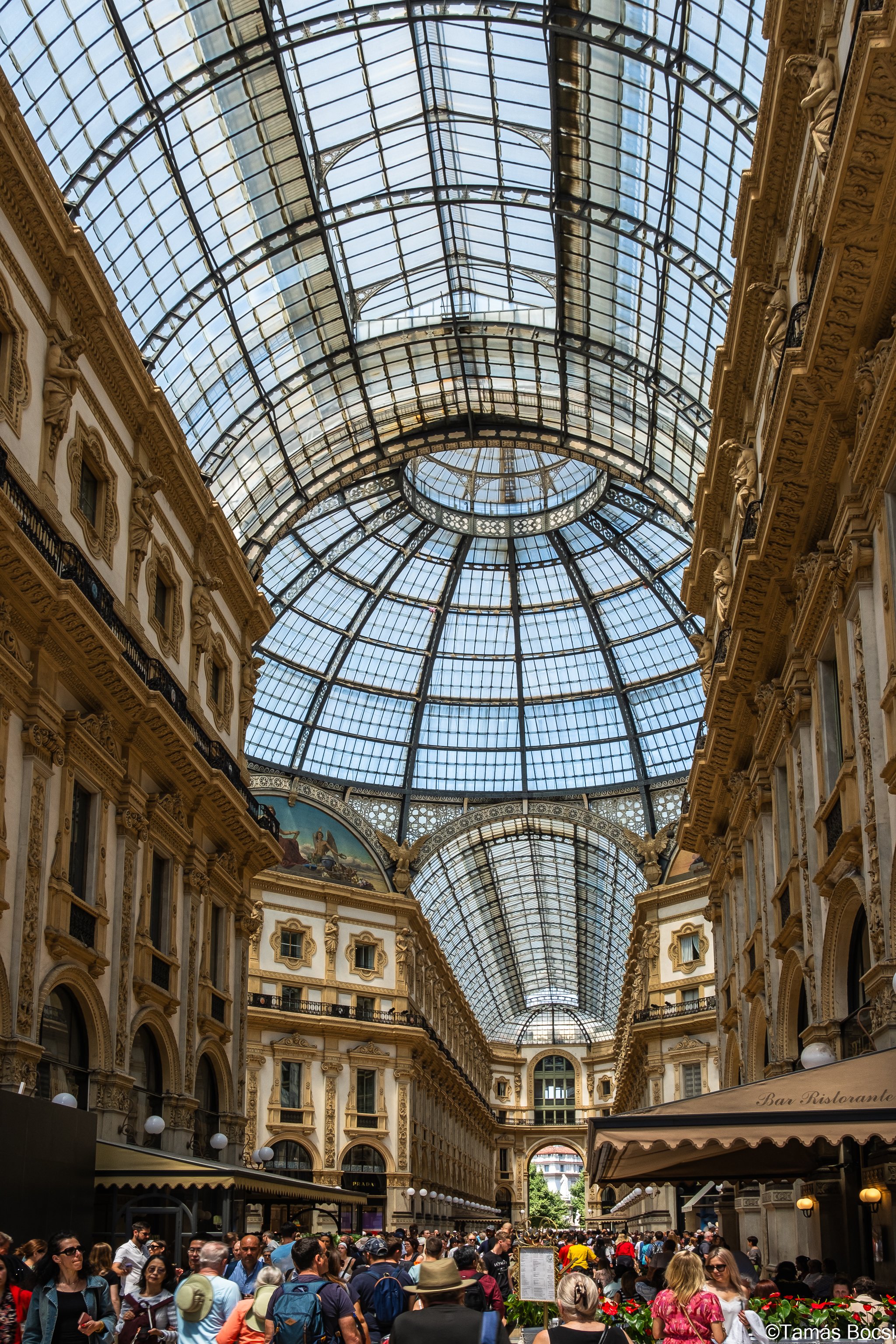 Interior of a grand shopping arcade with ornate architecture and a large glass dome ceiling, filled with many people shopping and walking.