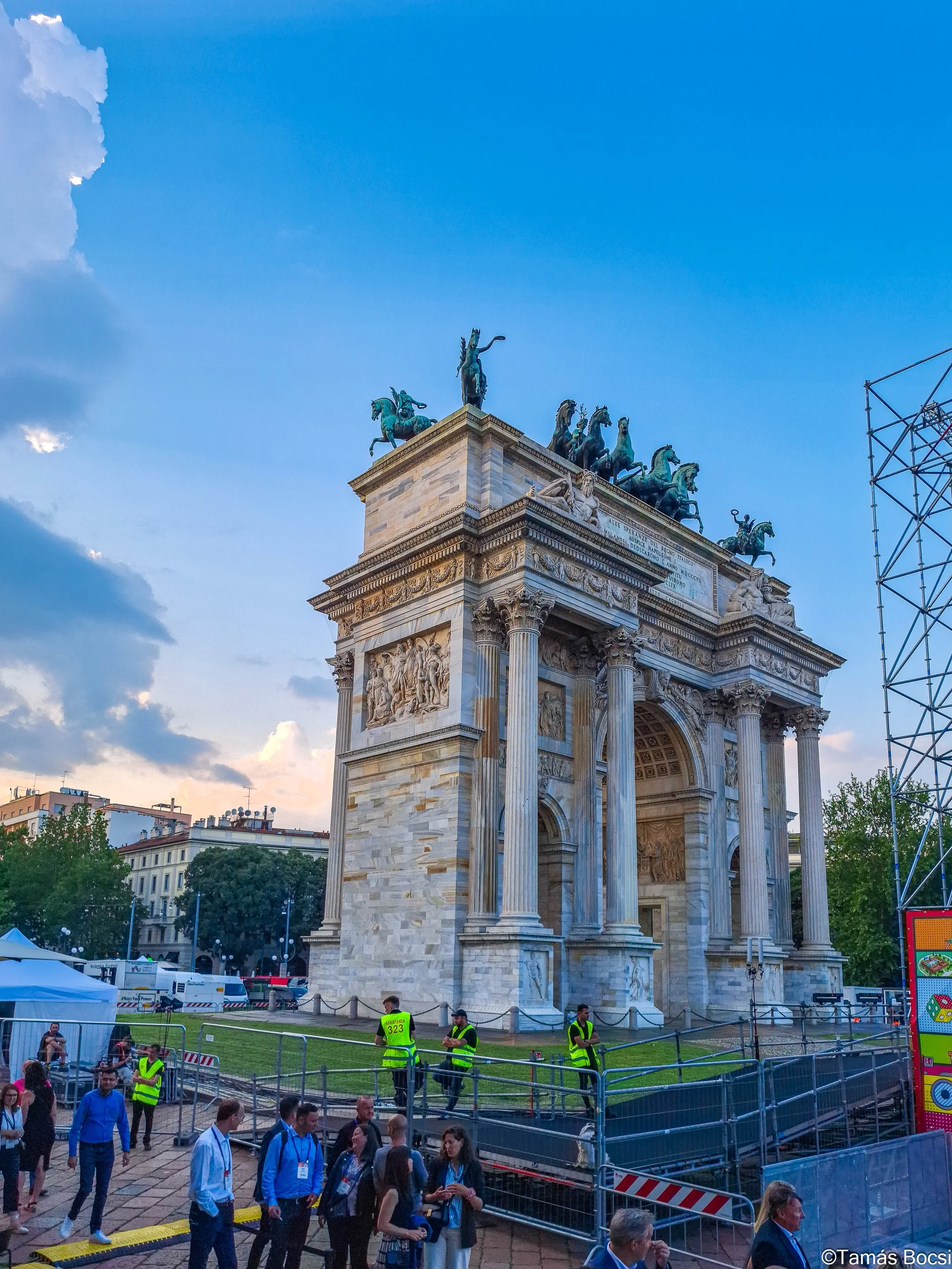 The Arc de Triomphe monument in Milan, Italy, with people and security personnel nearby, and a partly cloudy sky.