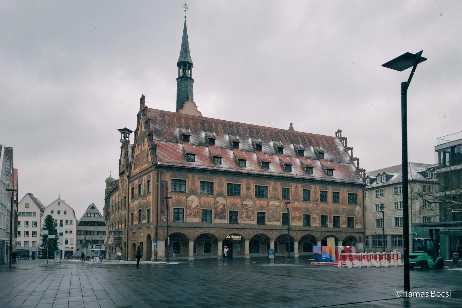 Historic building with a steep red roof, murals on its facade, and a pointed tower. The scene shows a wet, empty city square on a rainy day with palm trees and modern buildings in the background.