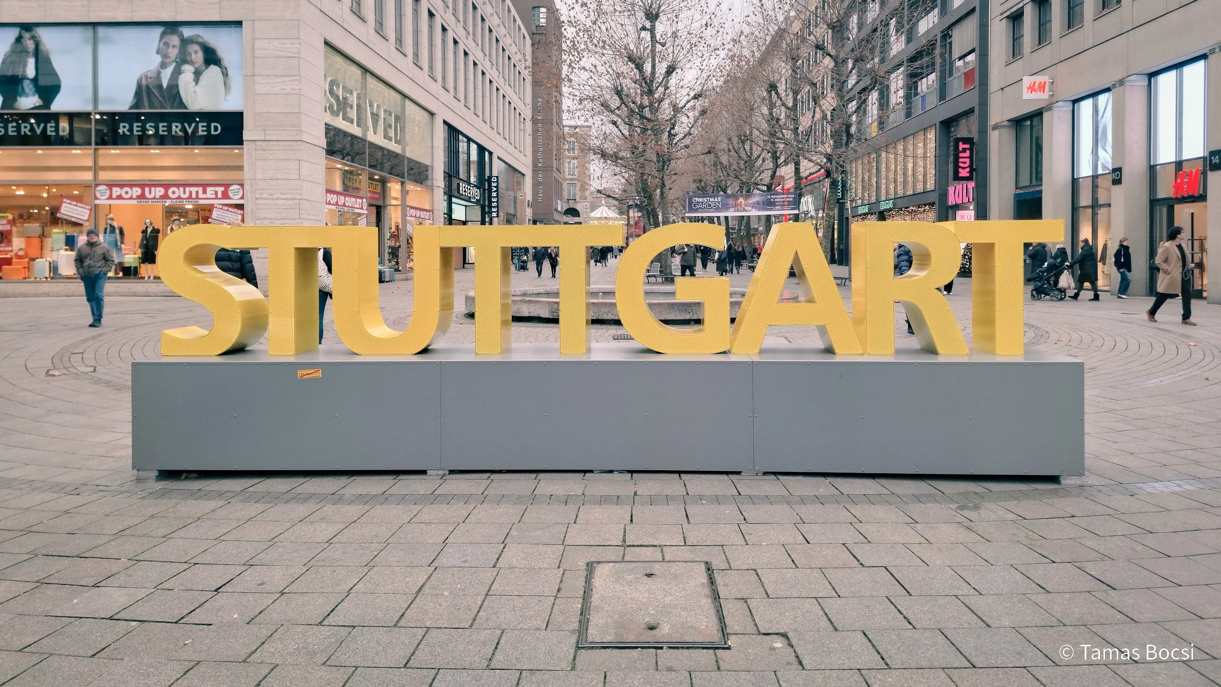 Large yellow letters spell out 'STUTTGART' on a grey platform in a pedestrian city street with stores and people.