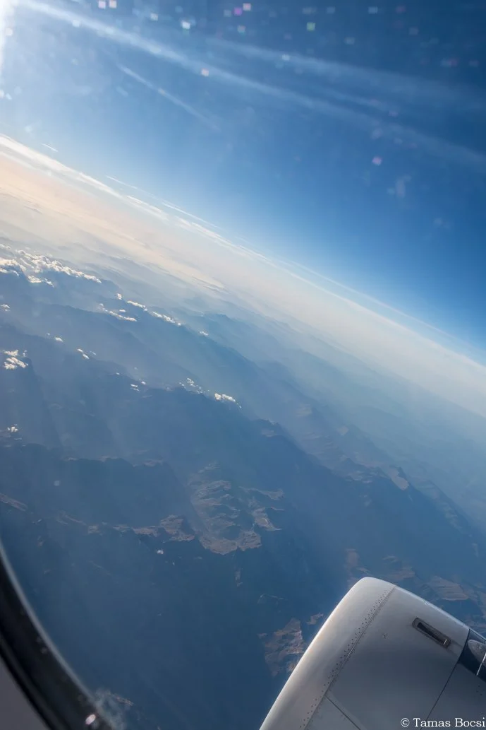 View from an airplane window showing the aircraft engine near the bottom right, with a landscape of mountains and clouds below, and a clear blue sky above.