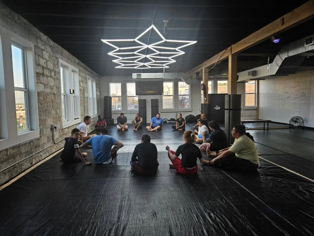Group of people sitting on the floor in a circle inside a fitness studio, with large windows, exposed brick walls, and a ceiling light fixture.