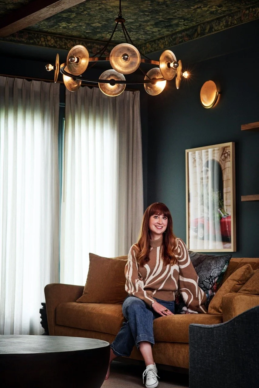 A woman with long red hair, wearing a tan and white patterned sweater and blue jeans, sitting on a brown couch in a warmly lit living room, with light-colored curtains behind her and a framed photo on the teal-colored wall.