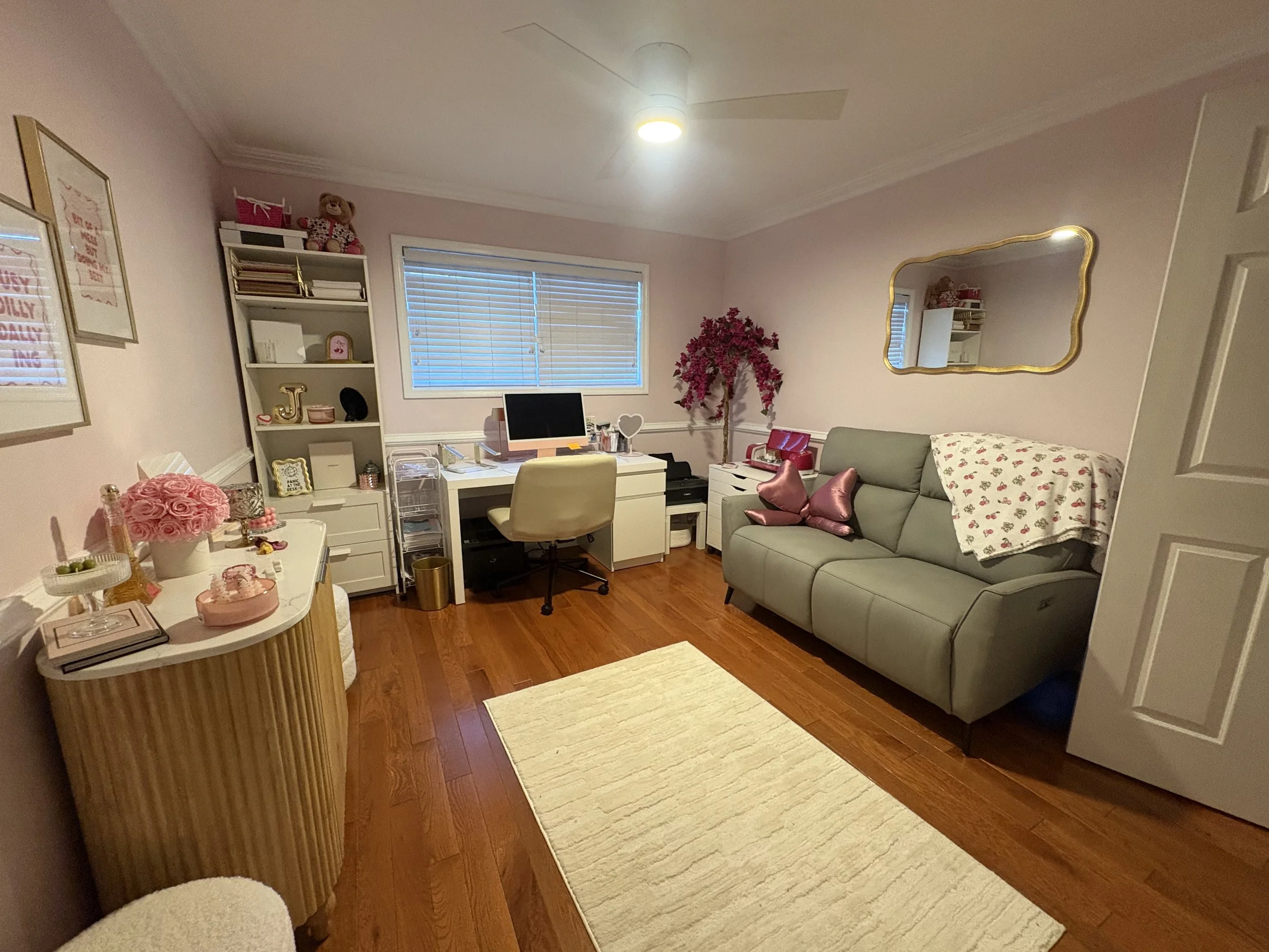 A cozy, well-decorated, well-organized, decluttered living room with pink walls and hardwood floors, featuring a computer desk with a beige chair, a gray sofa with pink pillows, a white side table with a large mirror on the wall.
