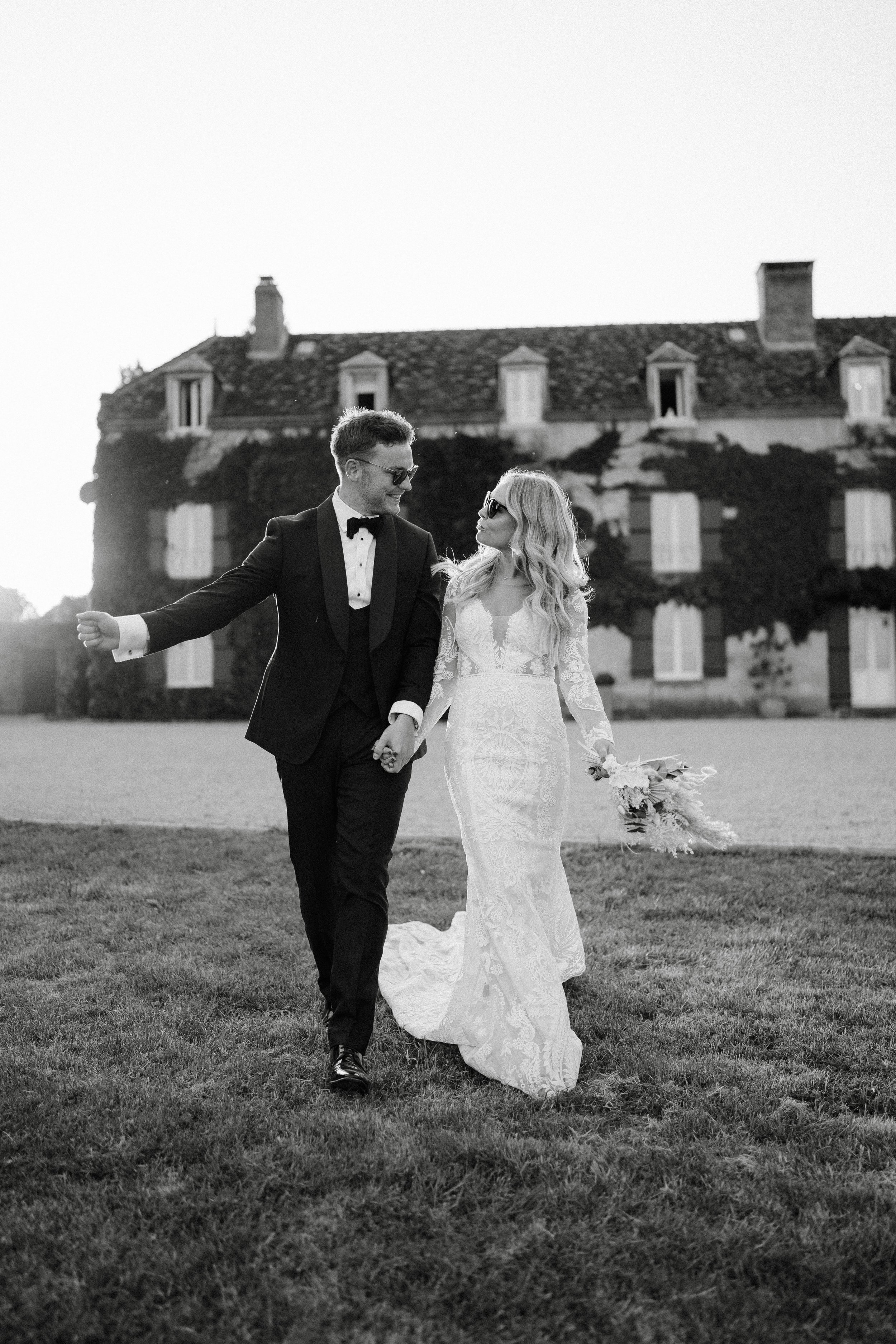 A black-and-white photo of a newlywed couple holding hands and walking on a lawn in front of a large house. The groom is dressed in a tuxedo and sunglasses, while the bride wears a lace wedding gown and holds a bouquet of flowers.
