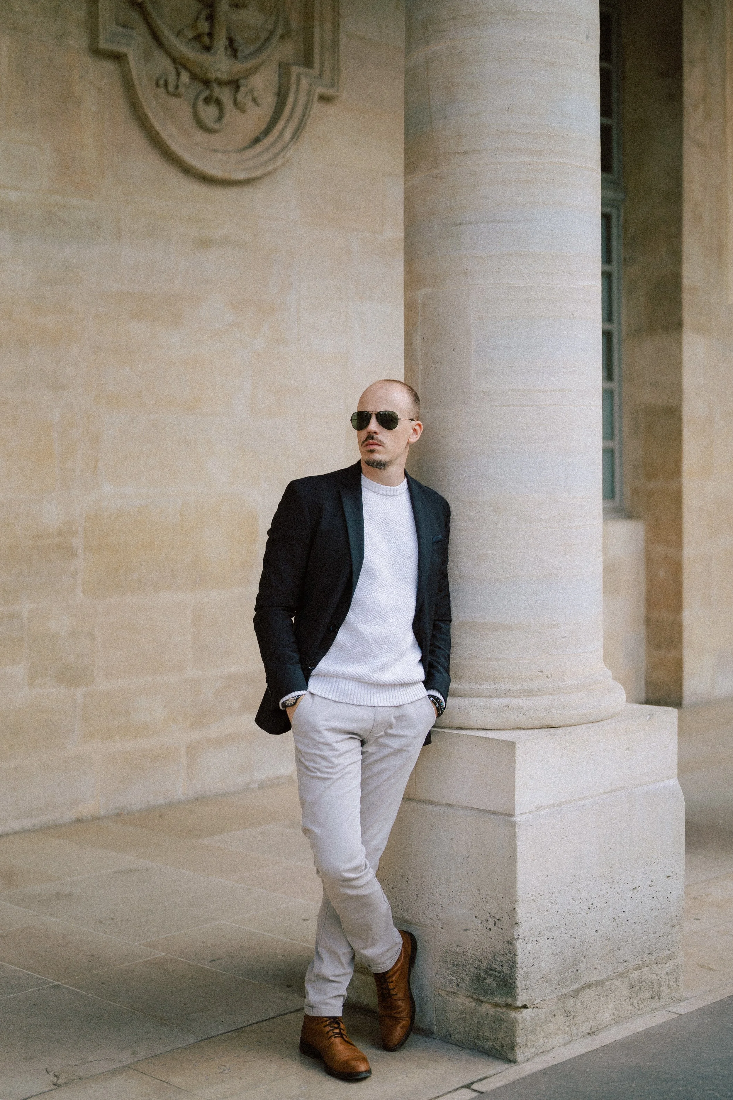 Vivien Malagnat, wedding photographer in sunglasses, a black blazer, white sweater, light gray pants, and brown boots stands against a large stone column outside a classical building in Paris