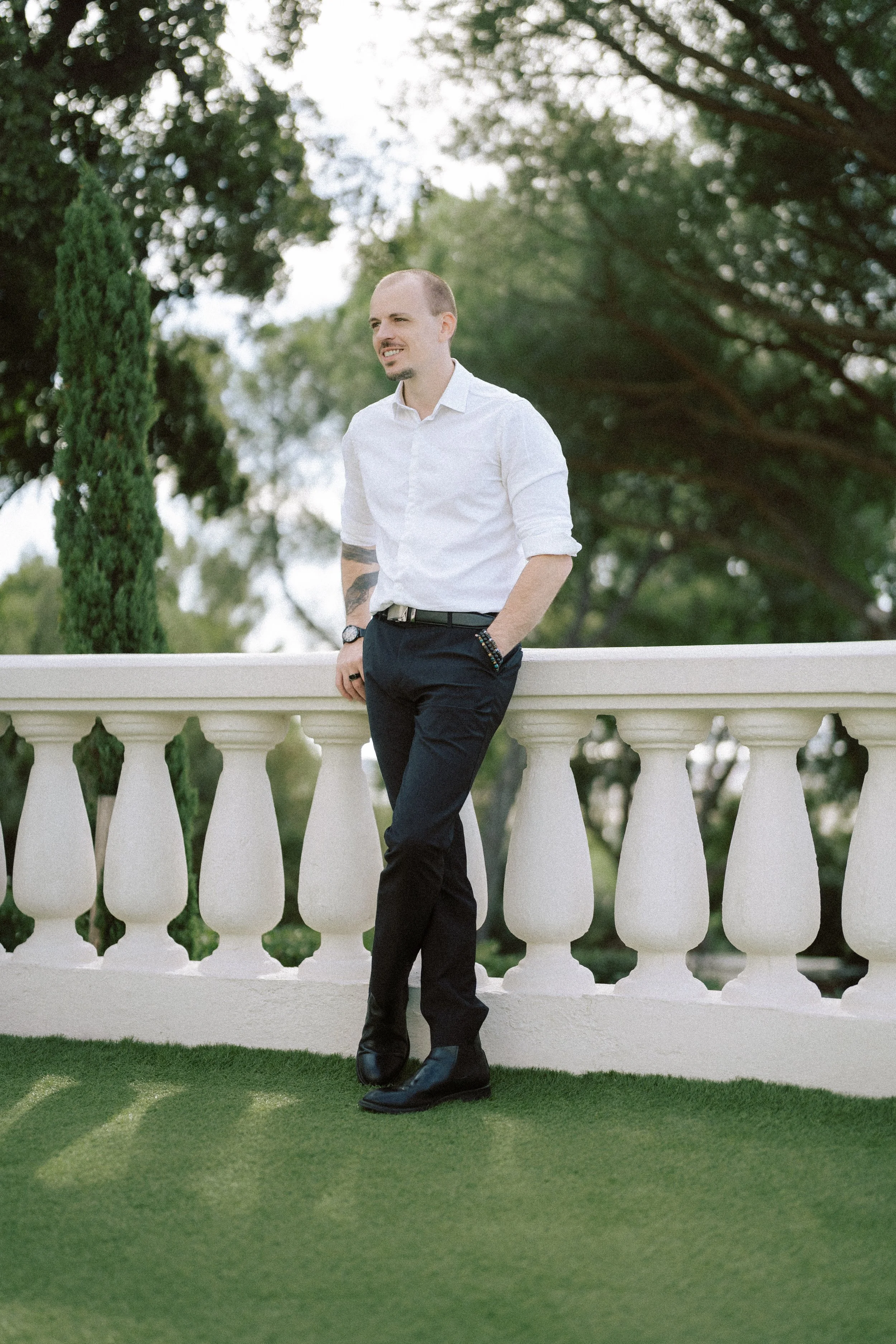 Vivien Malagnat, wedding photographer, dressed in a white shirt and dark pants leaning on a white balustrade outdoors on a grassy area, with trees and sky in the background at castel Bay.