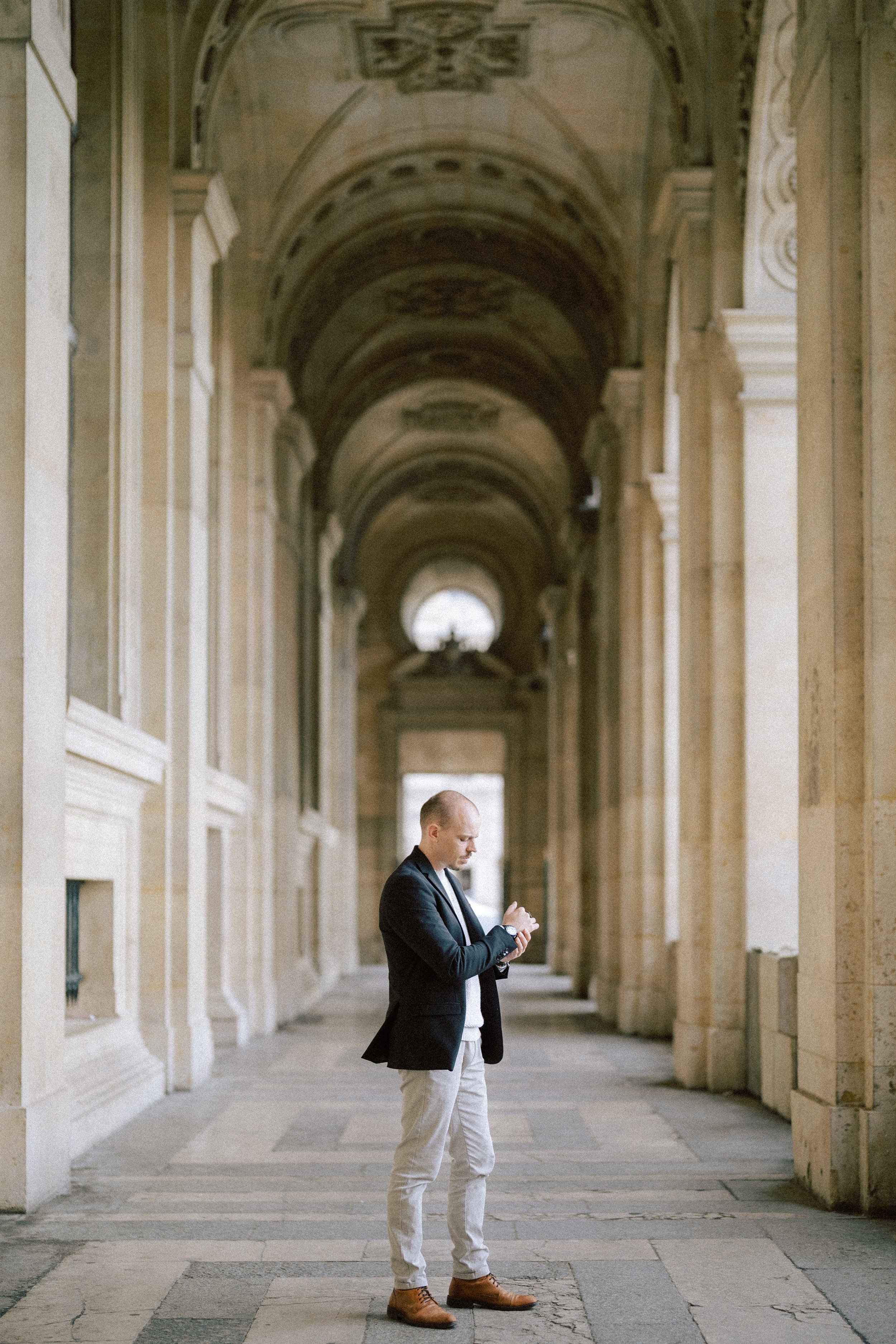 Vivien Malagnat, wedding photographer, in attire looking at his watch in a grand, arched, stone corridor with high ceilings and columns at Paris, near Louvre.