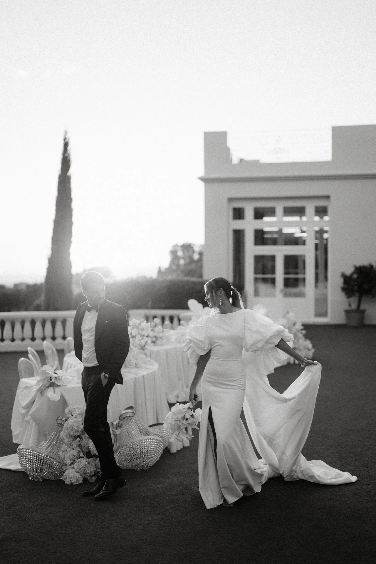 A black-and-white photo of a bride and groom at an outdoor wedding reception. The groom is in a tuxedo, standing with his hands in his pockets at castel Bay