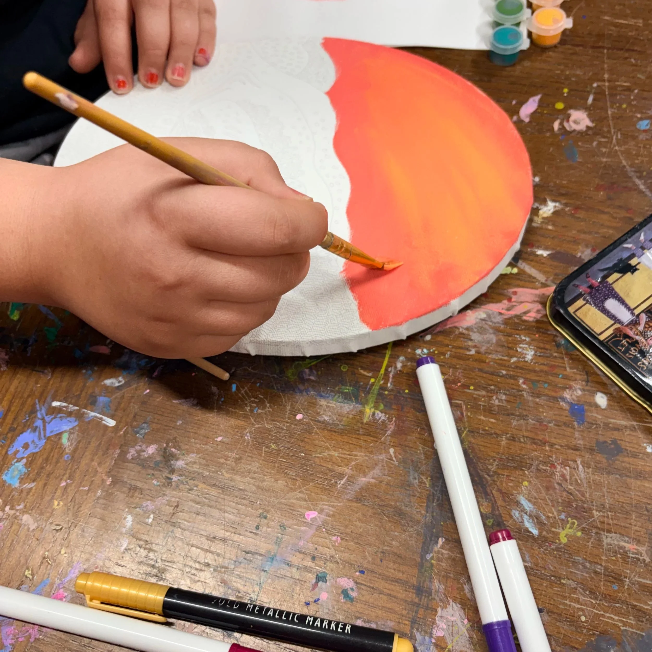 Children engaging in a multimedia art activity, using brushes and paint to create on paper, with various cups of paint and art supplies on the table.