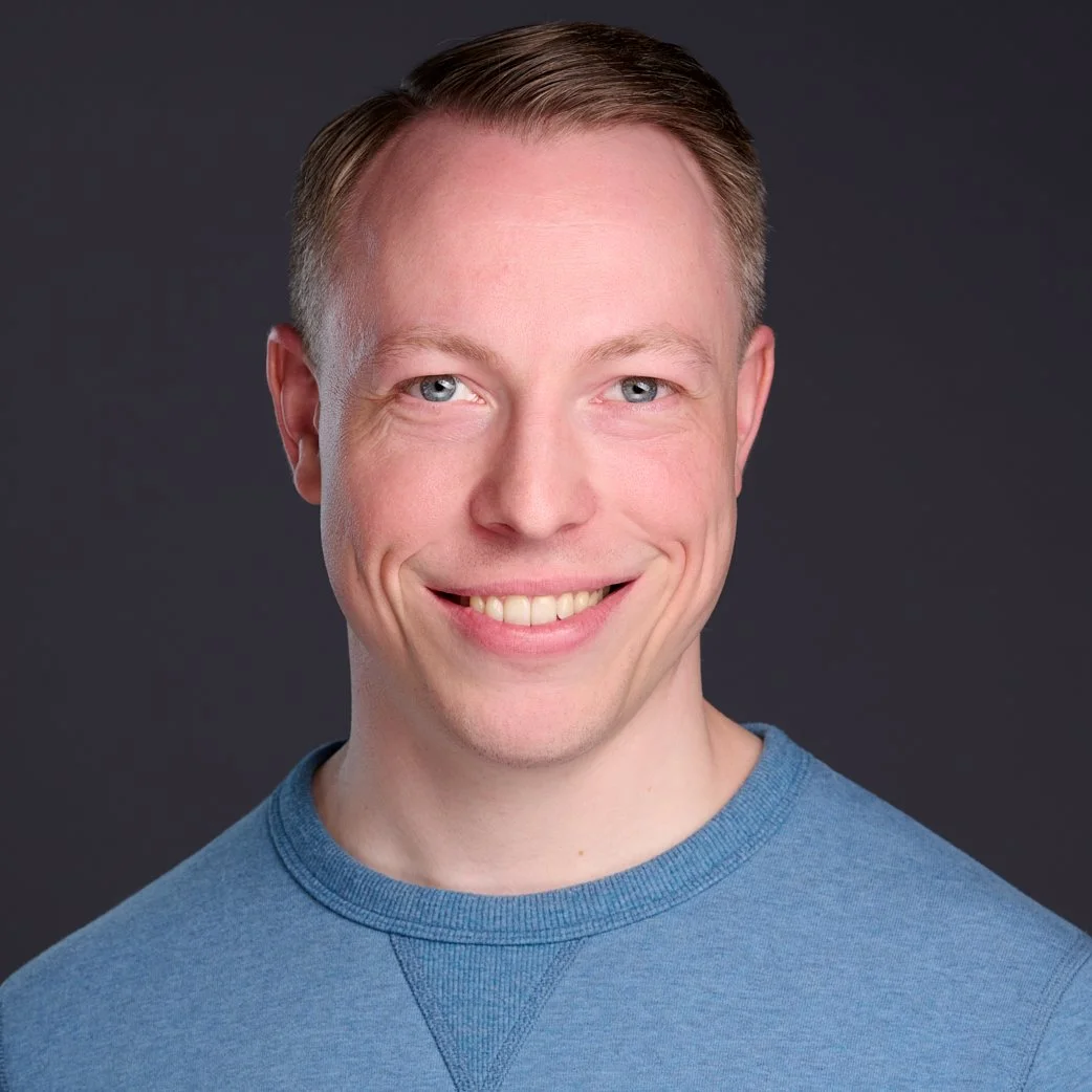 A smiling man with short light brown hair, blue eyes, and fair skin, wearing a blue crew neck shirt, against a dark background.