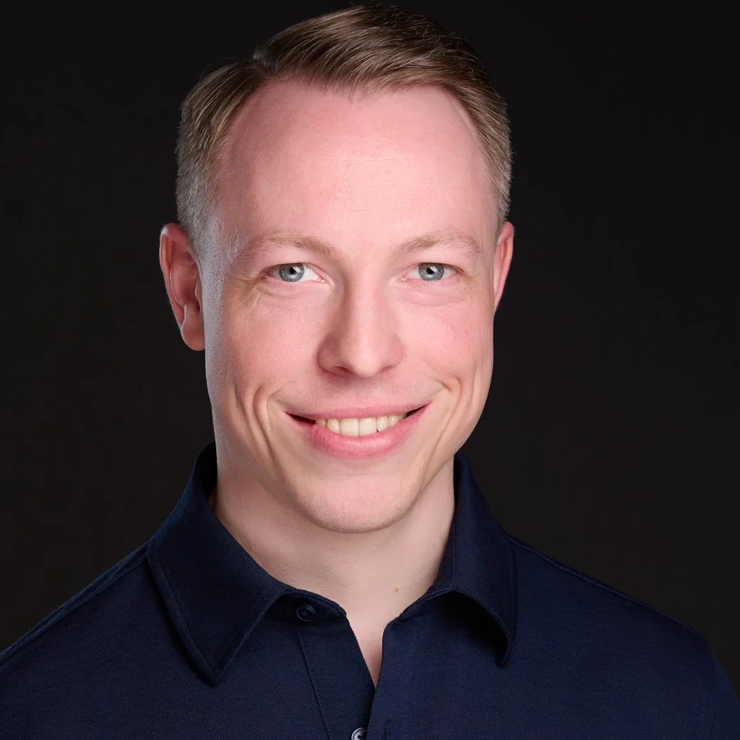 Portrait of a smiling young man with short blonde hair, blue eyes, wearing a dark navy collared shirt against a dark background.