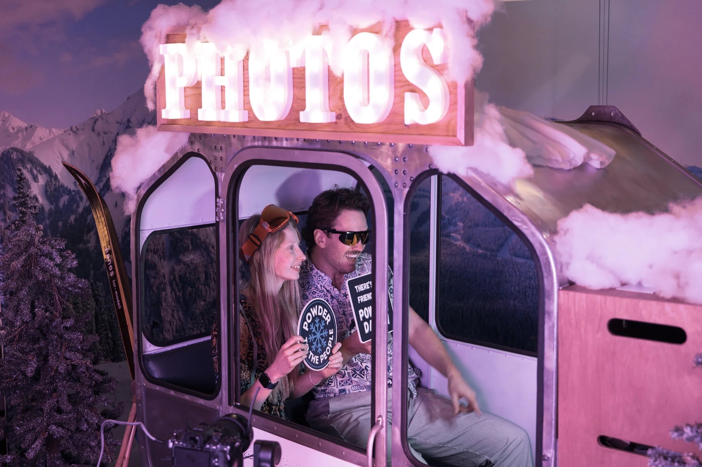 A couple inside a ski gondola with a sign saying 'PHOTOS' above them, holding photo booth signs, with snow-covered trees and mountain scenery in the background, pink and purple lighting.