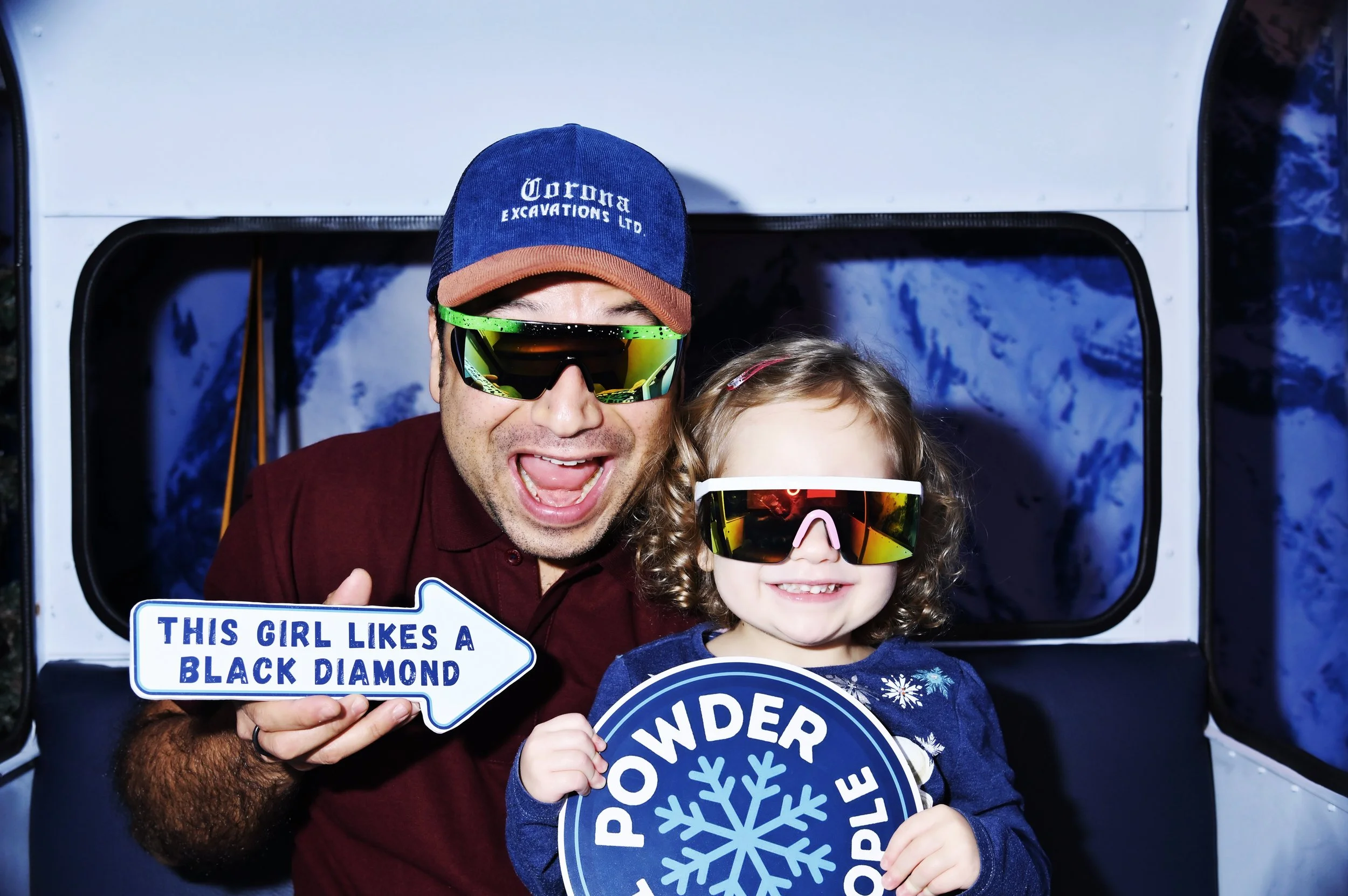 A smiling man and young girl in winter clothing and sunglasses pose for a photo. The man holds a sign that says 'This girl likes a black diamond,' and the girl holds a sign with a snowflake and the words 'Powder People.' They are inside or near a vehicle or snowmobile, with snow seen through the window in the background.