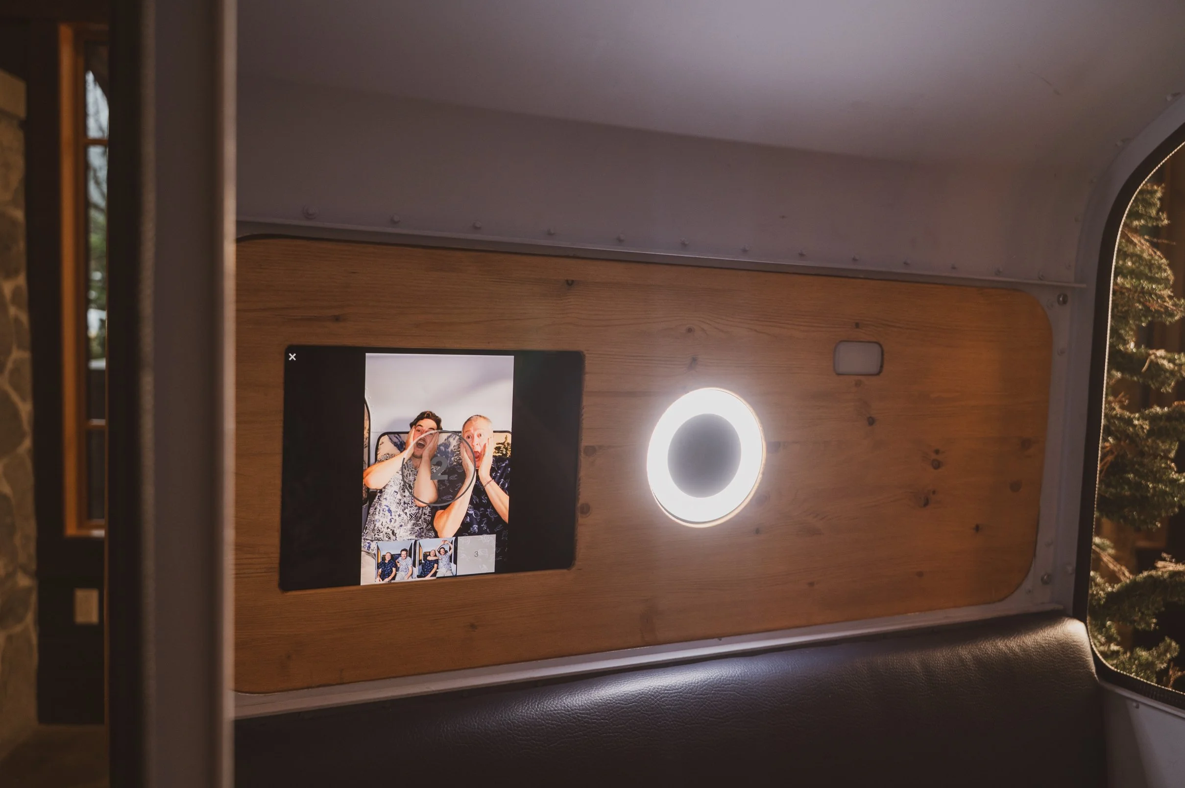 A photo booth inside a vehicle features a wooden panel with a digital screen showing two people taking a photo, a ring light, and a window with trees outside.