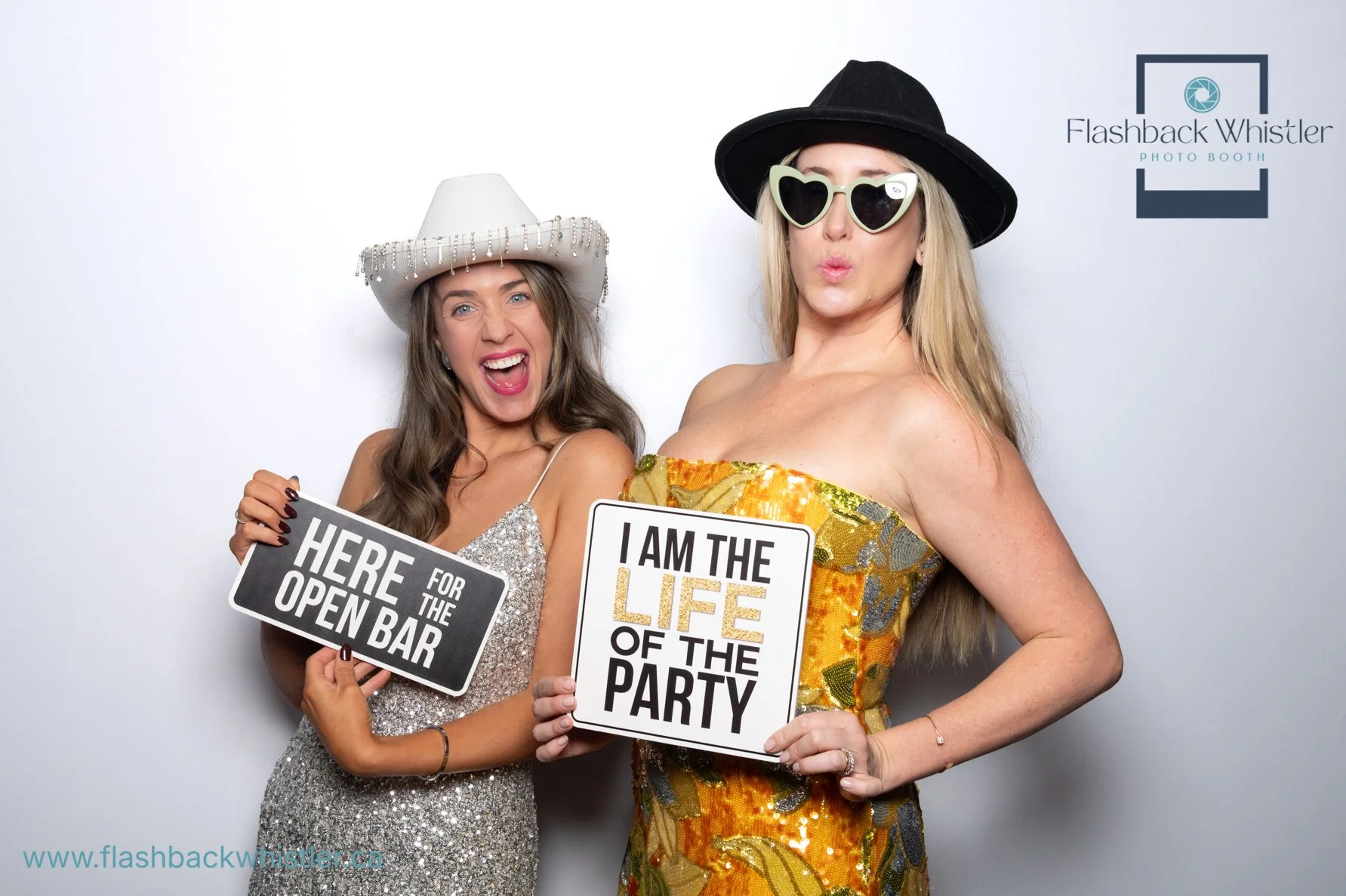 Two women dressed in party attire holding signs. One sign reads 'Here for the open bar' and the other reads 'I am the life of the party'. One woman wears a sparkly silver dress and a white cowboy hat with beads, and the other wears a strapless yellow dress with a black wide-brim hat and heart-shaped sunglasses. They are standing in front of a plain gray background with a logo that says 'Flashback Whistler Photo Booth' in the top right corner.