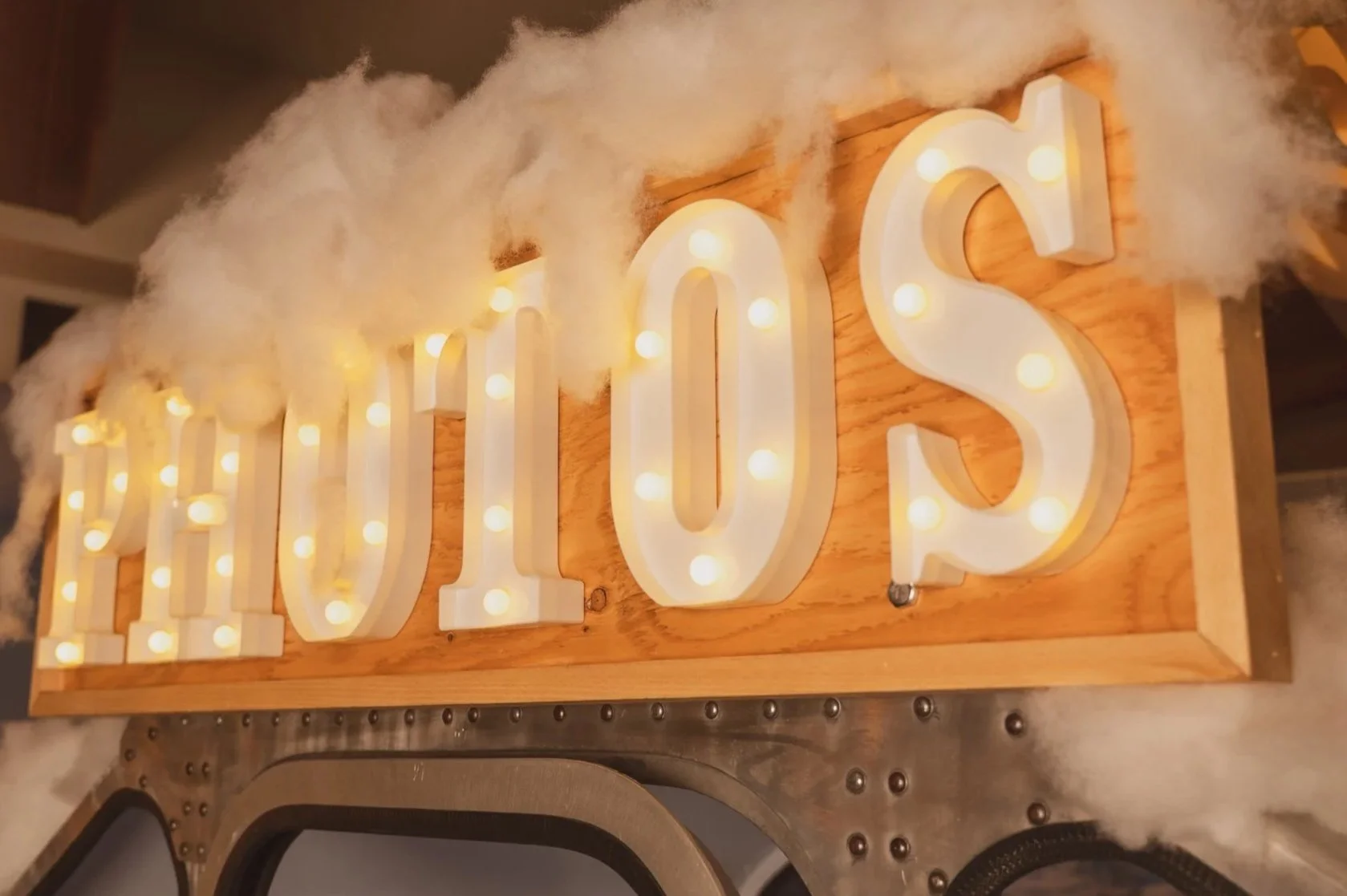 Decorative sign spelling 'HELLO' with illuminated marquee bulbs on a wooden background, topped with fluffy white material resembling clouds.