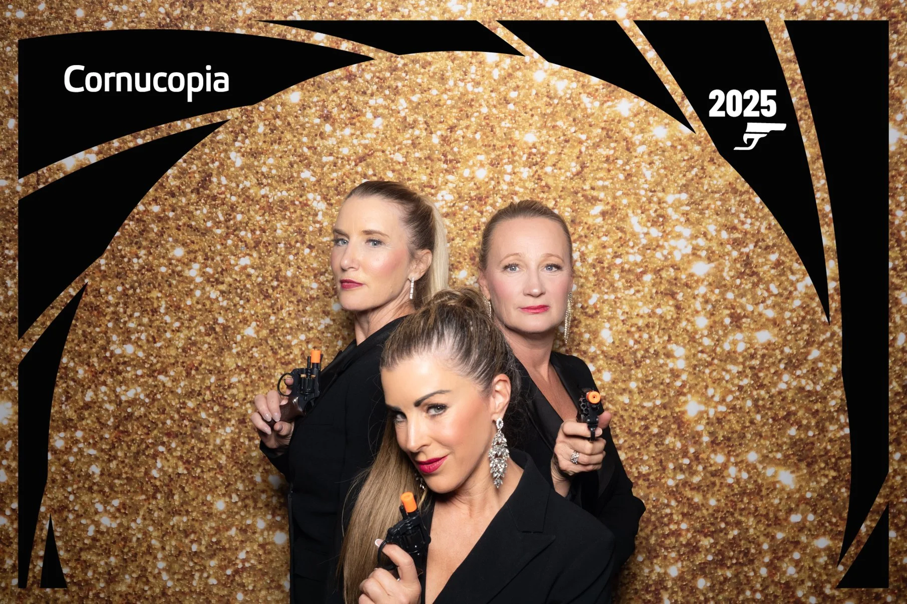 Three women in black outfits holding toy guns, posing in front of a gold glittery backdrop at a themed event with 'Cornucopia 2025' written on a black decorative frame.
