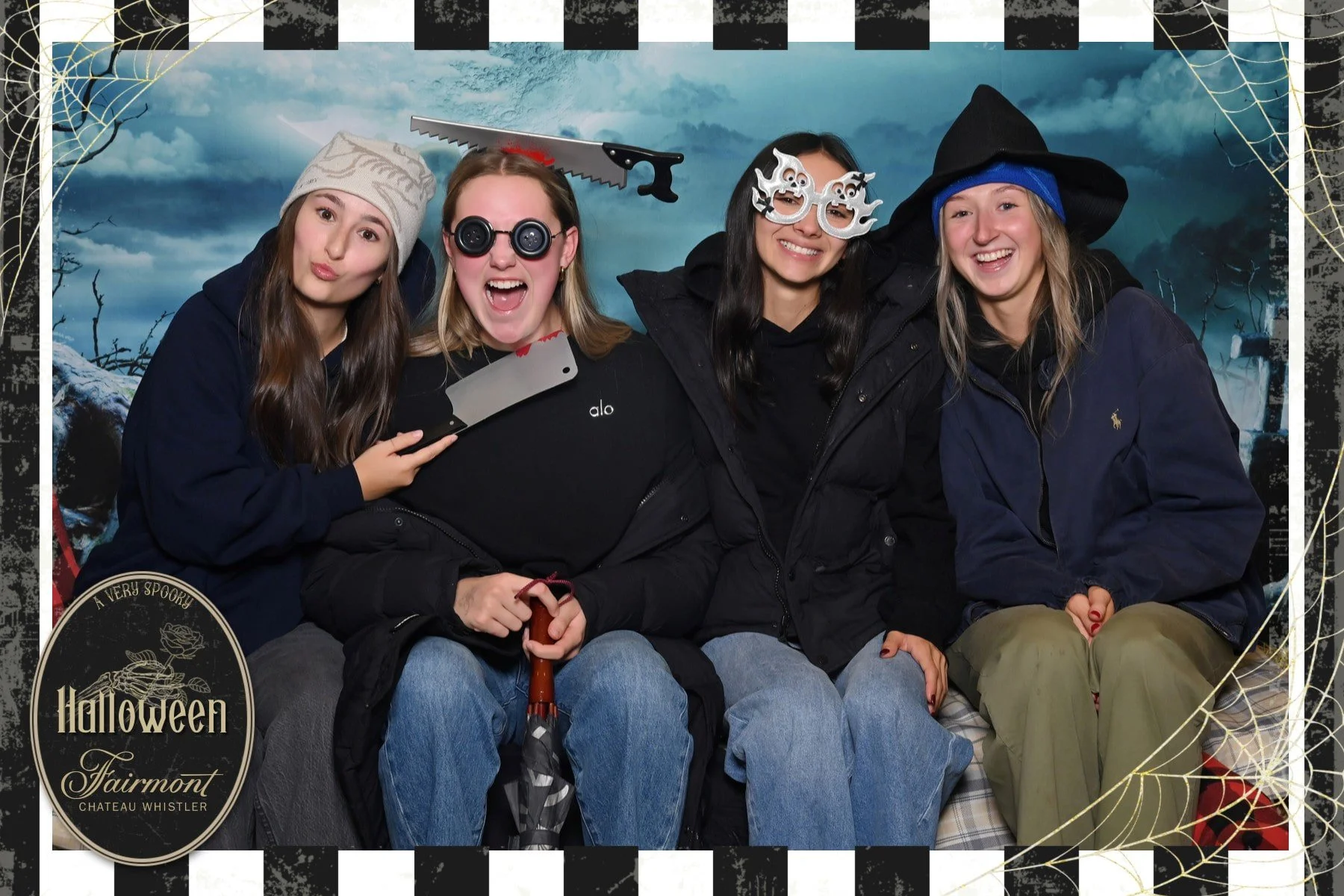 Four young women dressed in Halloween costumes and accessories, posing together for a photo with a Halloween-themed backdrop featuring a dark sky and spooky trees.