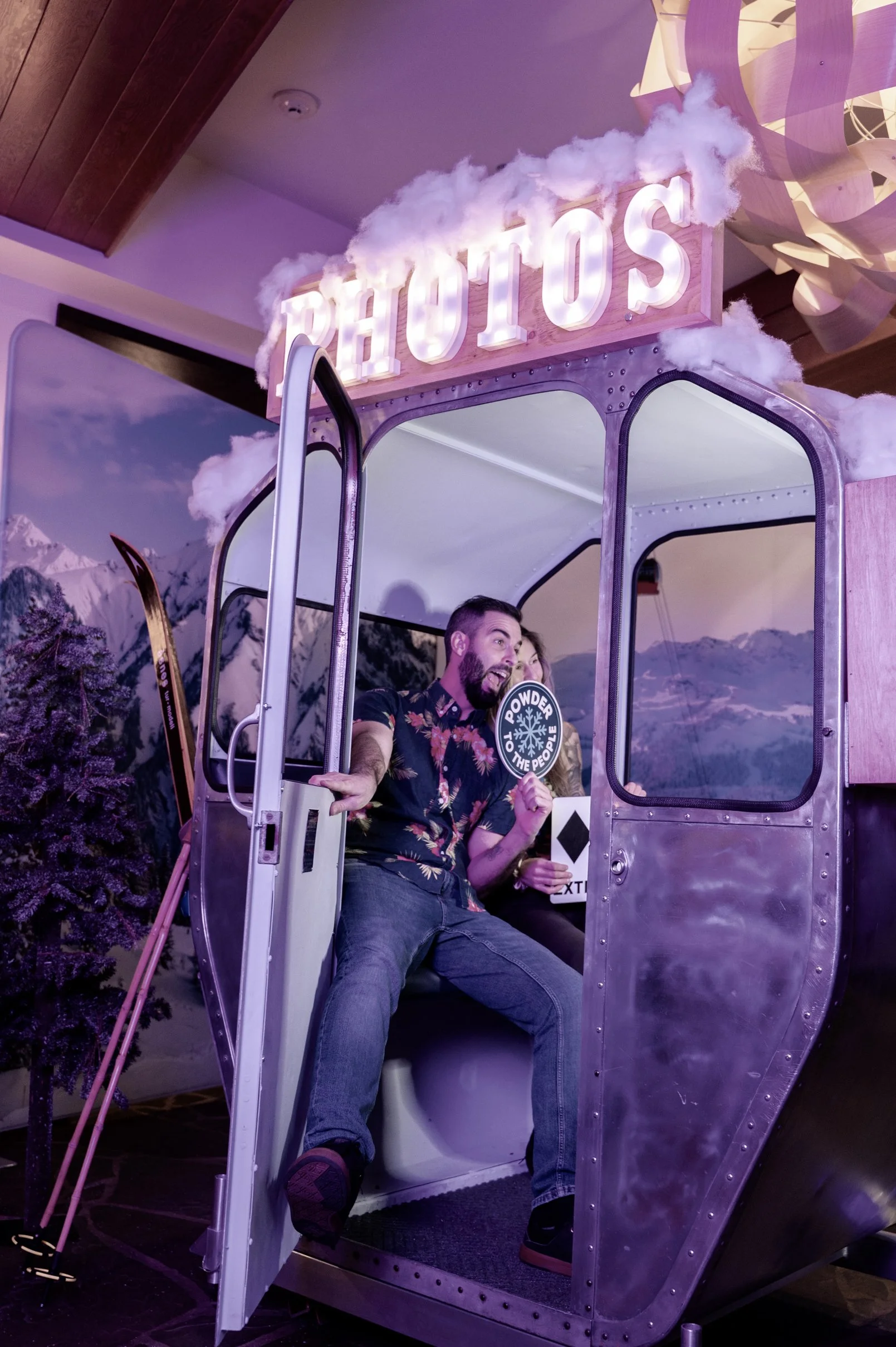 A man with a beard, wearing a Hawaiian shirt, sitting inside a metallic photo booth shaped like a ski gondola, holding a sign that says 'Powder to the People.' The photo booth has a fluffy, cloud-like decoration on top with a sign that reads 'PHOTOS.' In the background, there is a painted mountain scene and a pair of skis leaning against a tree.
