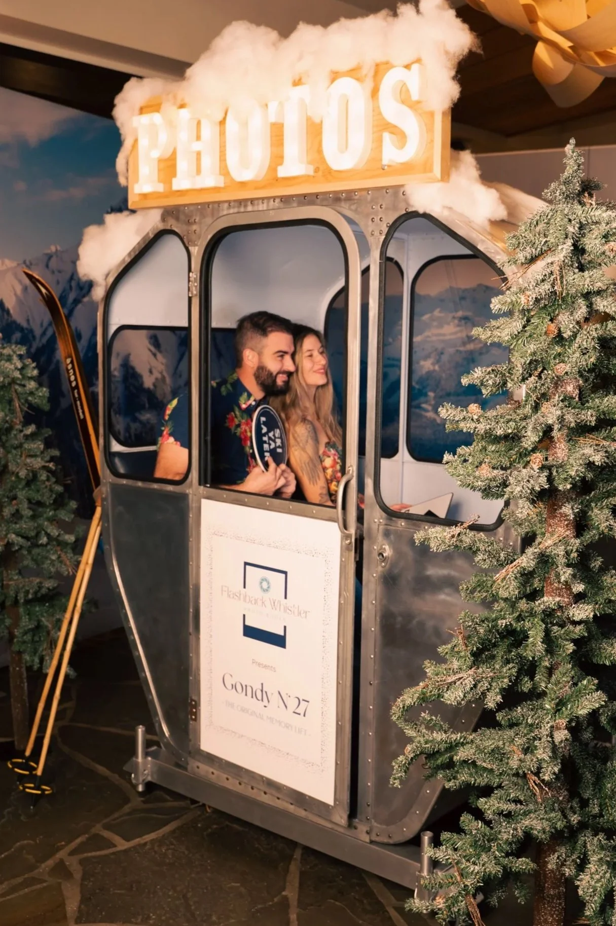 Two people sitting inside a small snow-themed cable car prop with a sign that reads "PHOTOS" on top, surrounded by decorated artificial snow-covered trees, with mountains and sky backdrop, and a sign on the cable car that says "Gondy N°27".