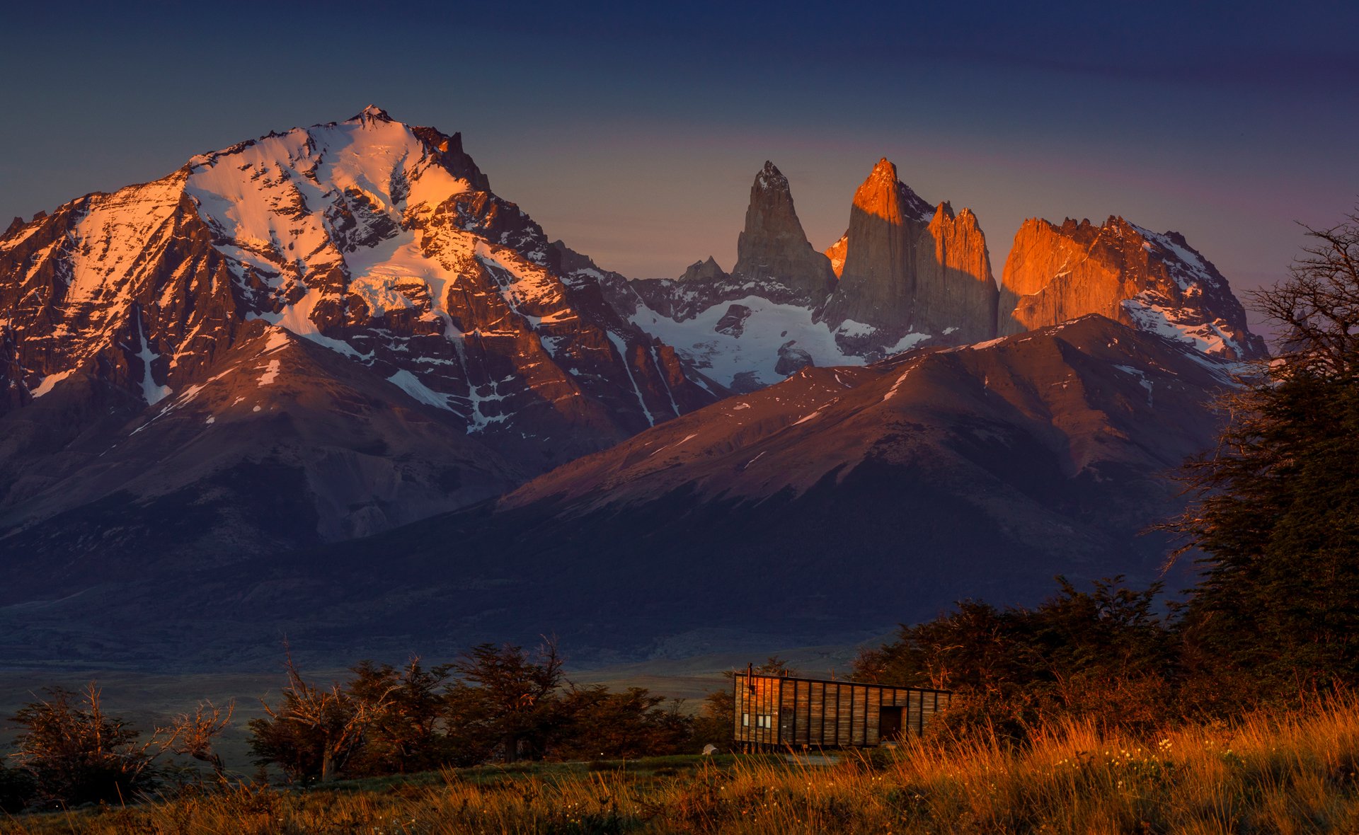 Montañas nevadas y rocosas al atardecer, con un paisaje de campo y árboles en primer plano.
