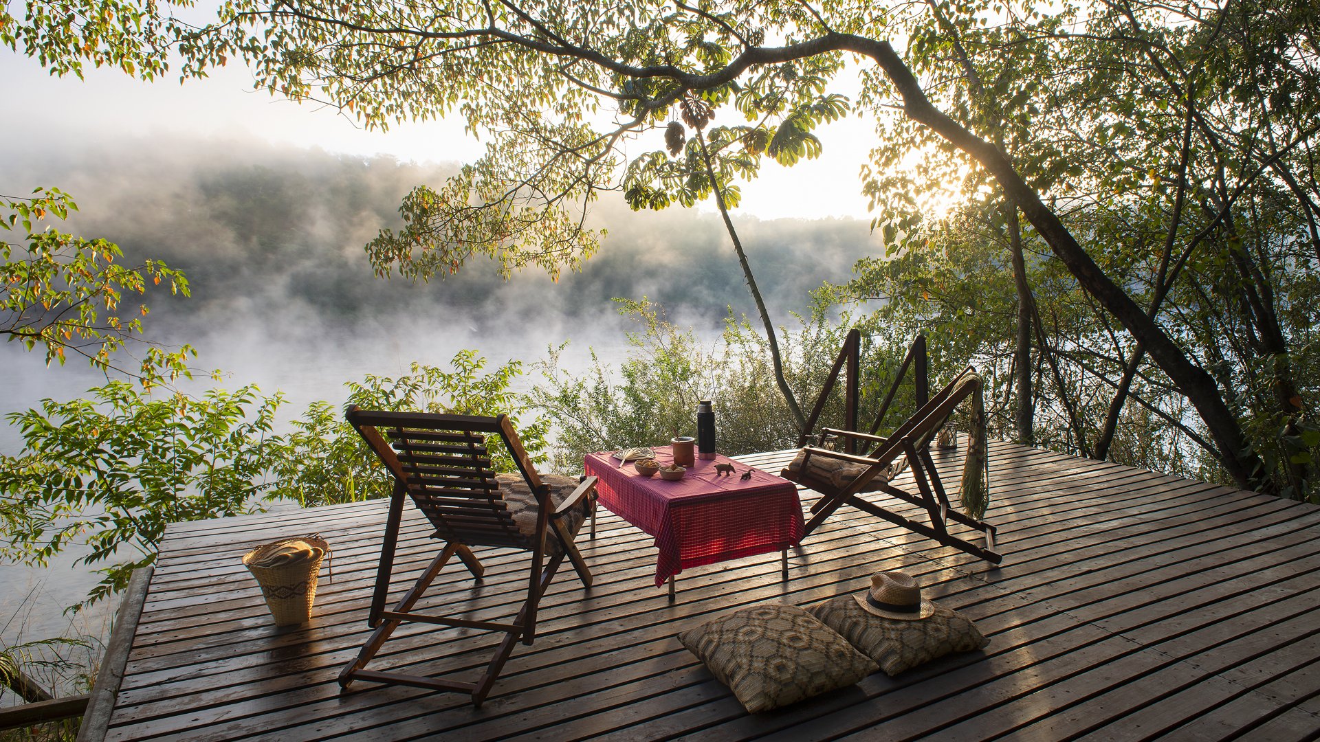 Una terraza de madera con sillones, una mesa con una mesa de playa y bebidas, colchonetas y sombrero, rodeada de árboles y vista a un río con niebla matutina.