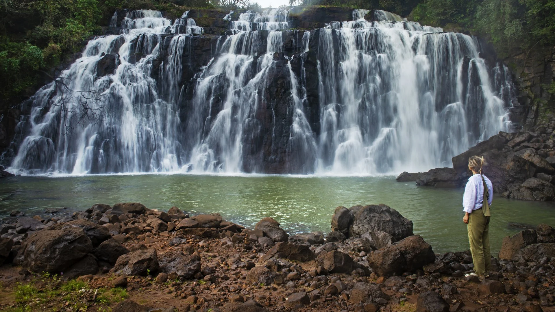 Una mujer con ropa clara y pantalones verdes mira una cascada en un paisaje natural rocoso con vegetación.