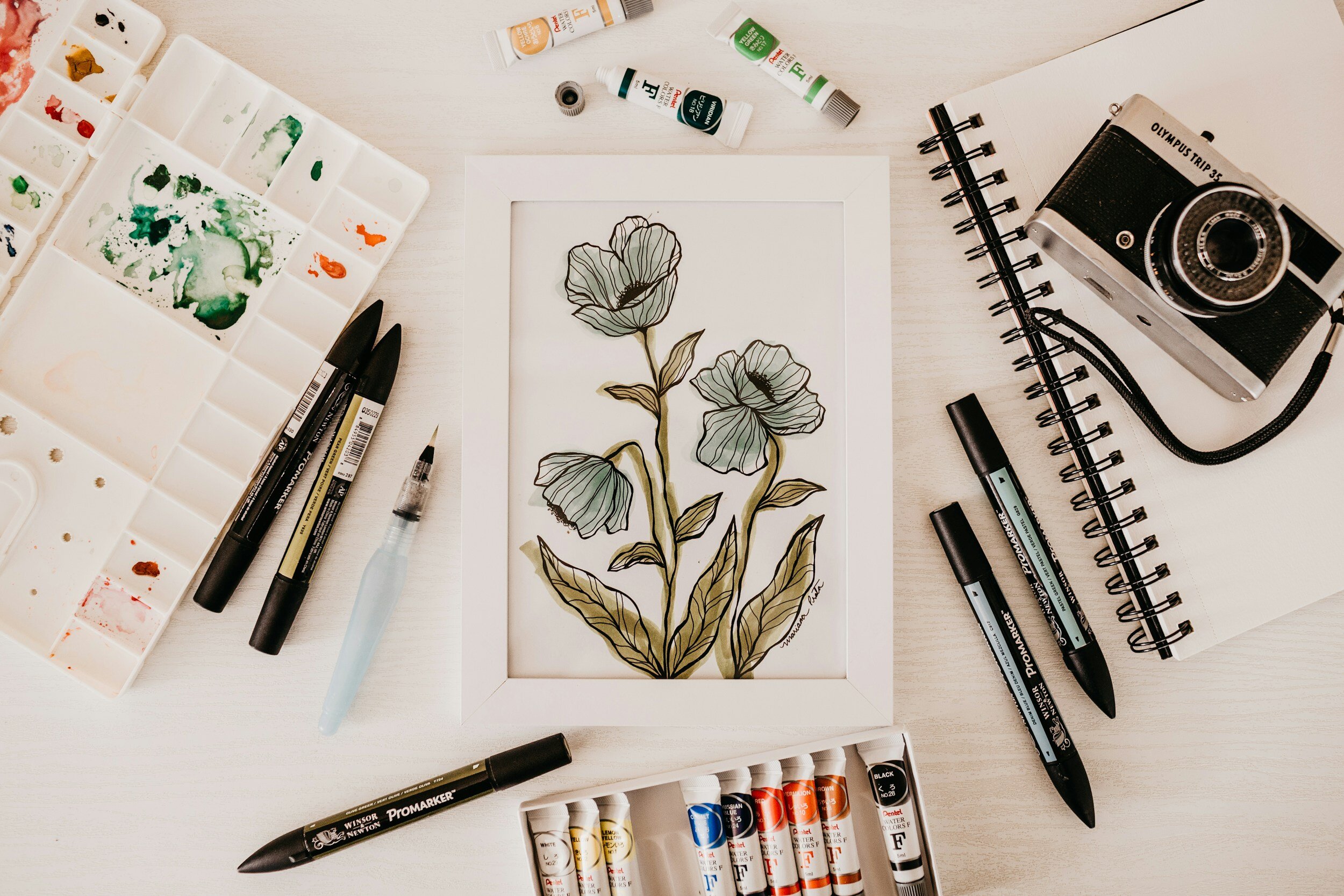 Art supplies and camera surrounding a botanical illustration of leaves and flowers in a white frame on a light-colored table.