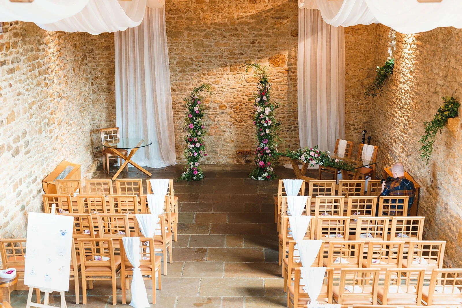A wedding ceremony taking place in a rustic, indoor venue with brick walls and wooden beams. The bride and groom are holding hands and smiling at each other. The bride is wearing a white wedding dress, and the groom is in a gray suit. Guests are seated, including a woman in a navy dress and large hat, and an older woman in a polka-dot dress. Floral decorations are visible at the front.