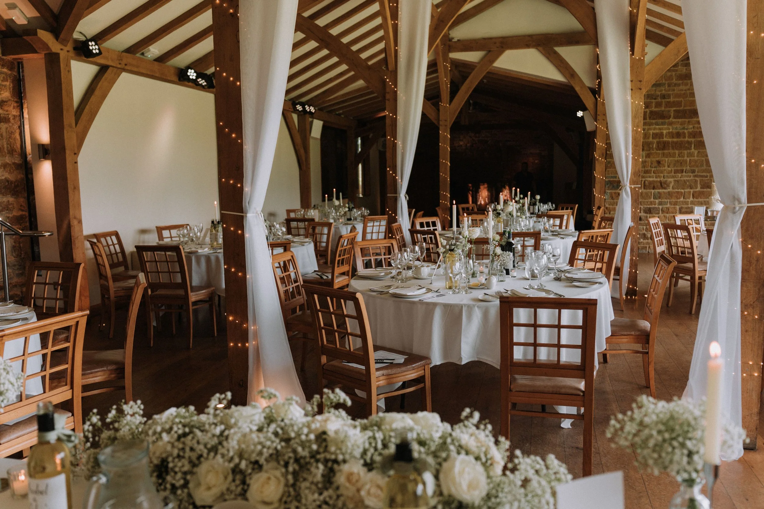 Wedding reception setup inside a rustic wooden venue with round tables dressed in white tablecloths, glassware, and floral centerpieces, decorated with fairy lights and white drapes hanging from wooden beams.