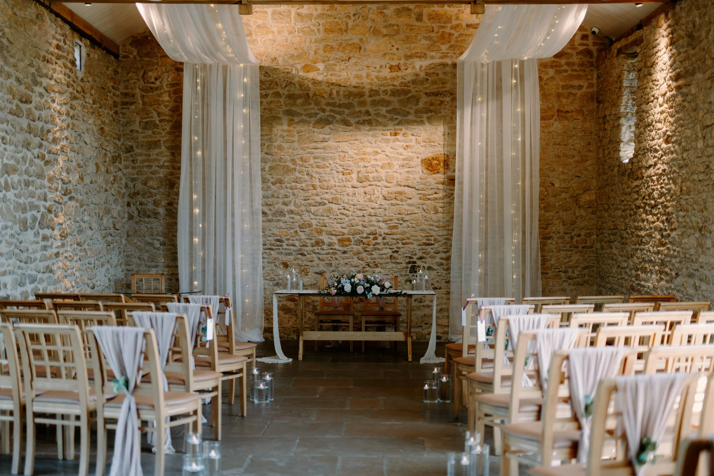 Indoor wedding ceremony setup with wooden chairs, floral arrangements in white vases, and white drapery on a wooden-beamed ceiling. Stone walls and large windows allow natural light.