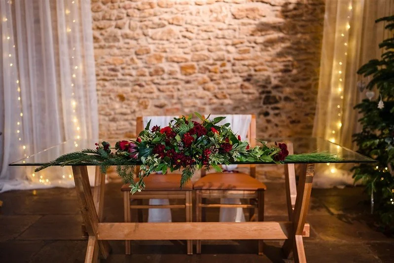 Elegant dining table set with white tablecloth, multiple wine glasses, white cups, and silverware, decorated with a tall candelabrum with lit candles and surrounded by wooden chairs. A blurred floral centerpiece is in the foreground. The room has subdued warm lighting.