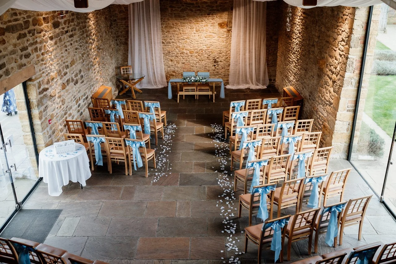 Elegant wedding reception with round tables, white tablecloths, floral centerpieces, glassware, and draped white fabric with fairy lights hanging from a wooden beam ceiling.