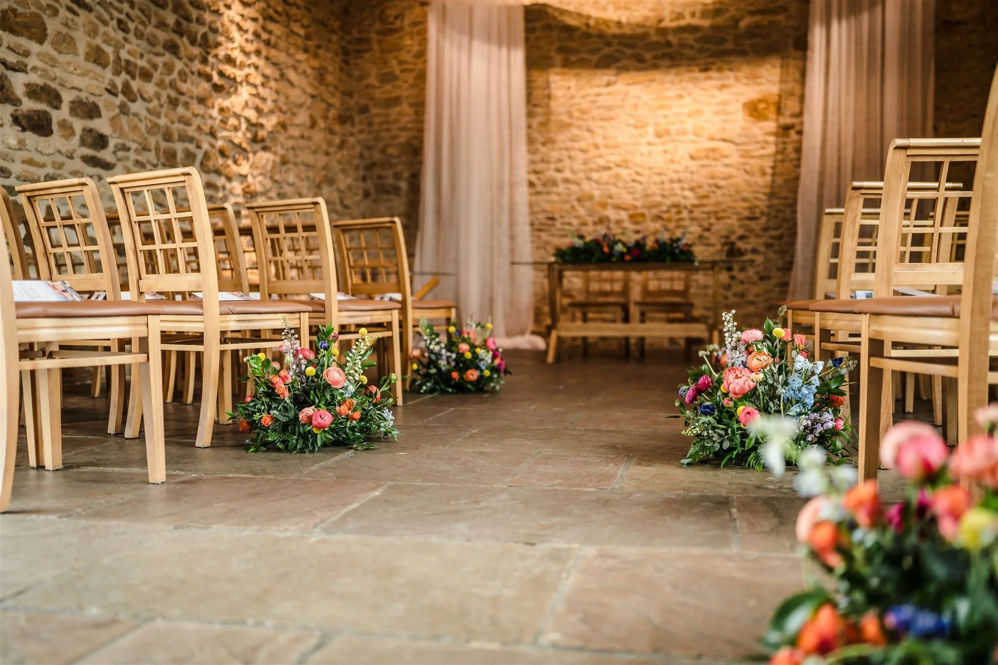 A floral centerpiece with pink roses, white flowers, and green leaves on a table, with a stone wall and a chandelier in the background.