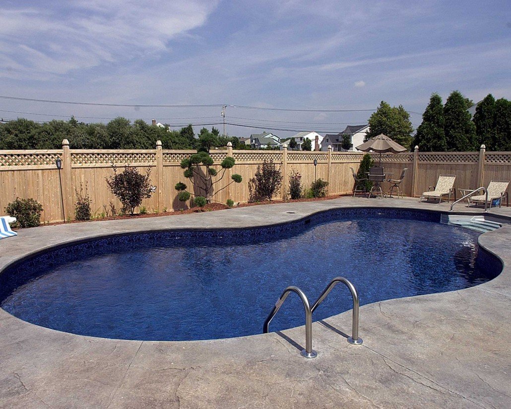 A backyard with a kidney-shaped swimming pool, surrounded by a concrete deck, a wooden privacy fence, and outdoor furniture including lounge chairs, a table with an umbrella, and a grilling area. Small trees and bushes are along the fence, and residential houses are visible in the background under a partly cloudy sky.