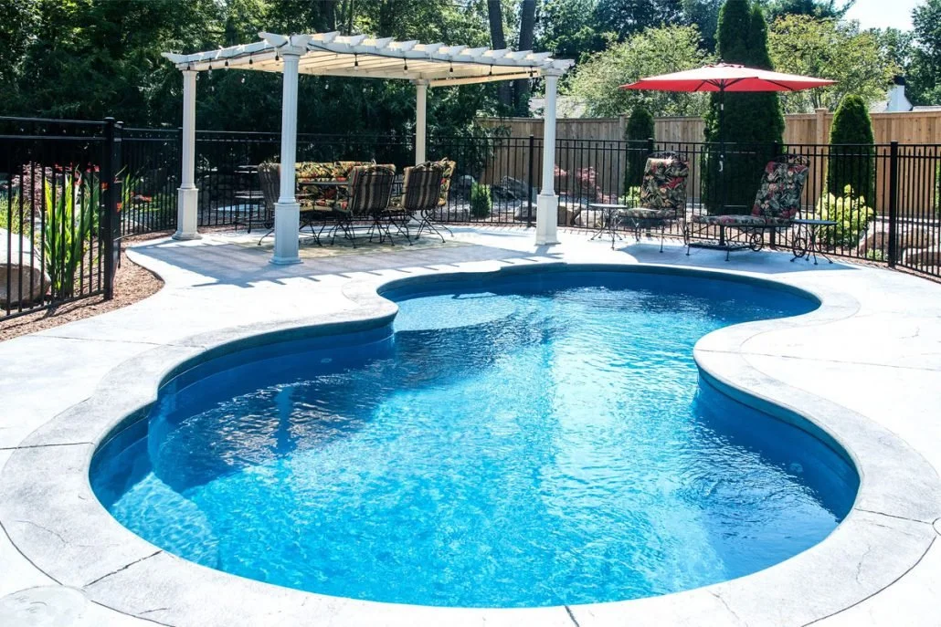 A backyard swimming pool with a freeform shape, surrounded by a concrete deck, a black metal fence, and outdoor furniture including a table with chairs, lounge chairs with floral cushions, an umbrella, and a white pergola with a canopy.
