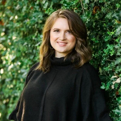 A woman with shoulder-length light brown hair wearing a black turtleneck sweater, standing outdoors near green leafy bushes, smiling at the camera.