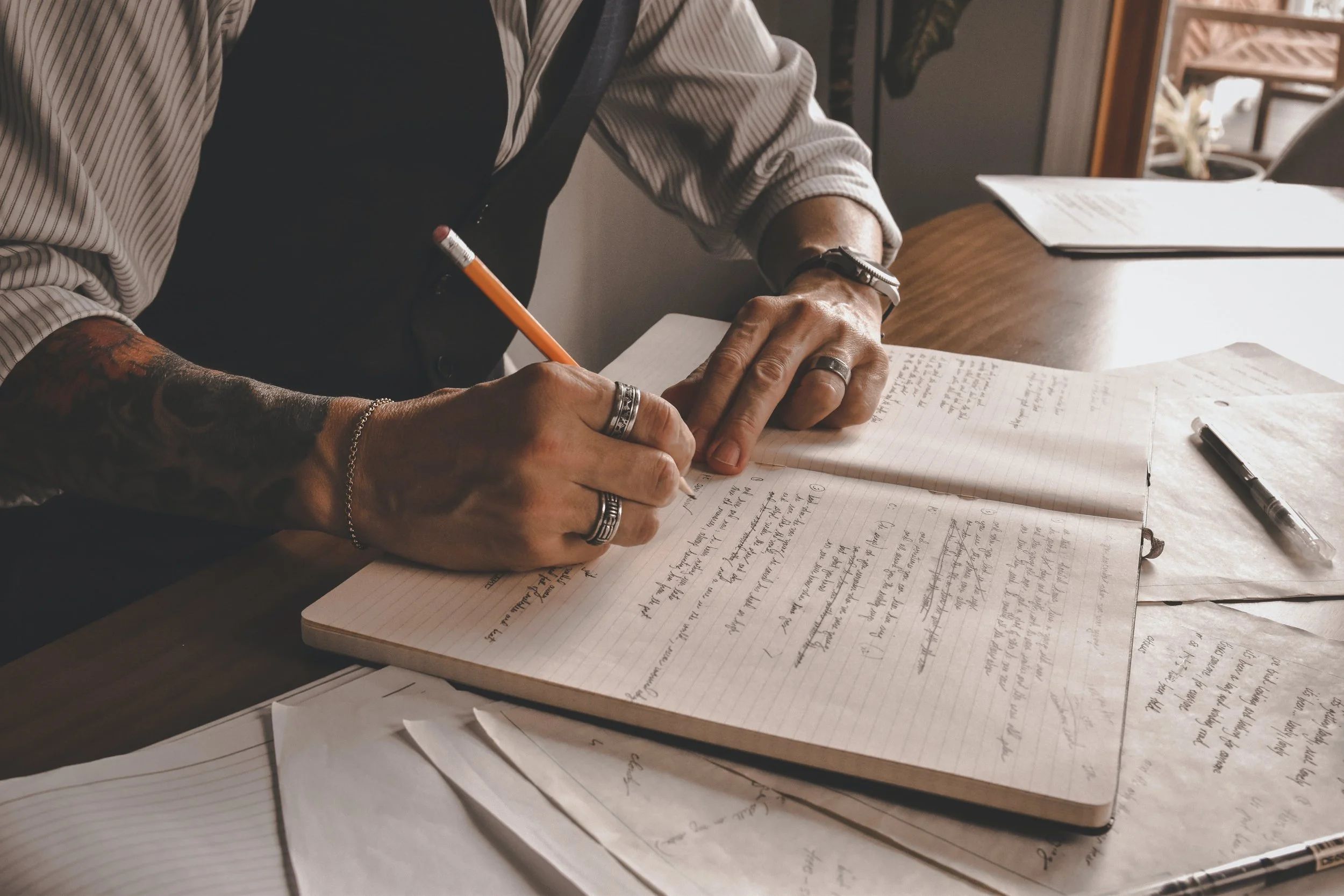 man with tatoos writing in a notebook with a pencil at a desk