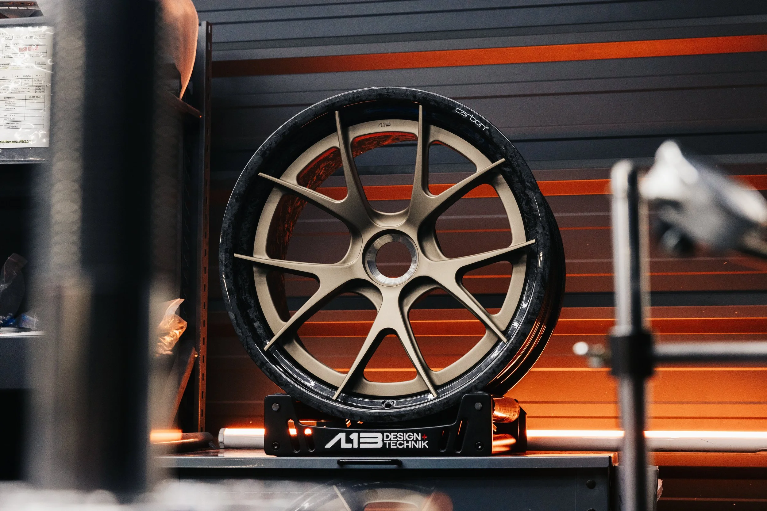 AL13 CF-G01 Carbon Series Wheel with a bronze split-spoke center and forged carbon fiber barrel, displayed on an AL13 Design + Technik stand in a workshop.