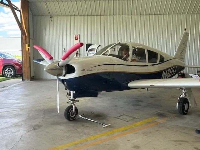 Small private aircraft inside a hangar, with a beige and blue exterior, pink covers on the propellers, and a person sitting inside.