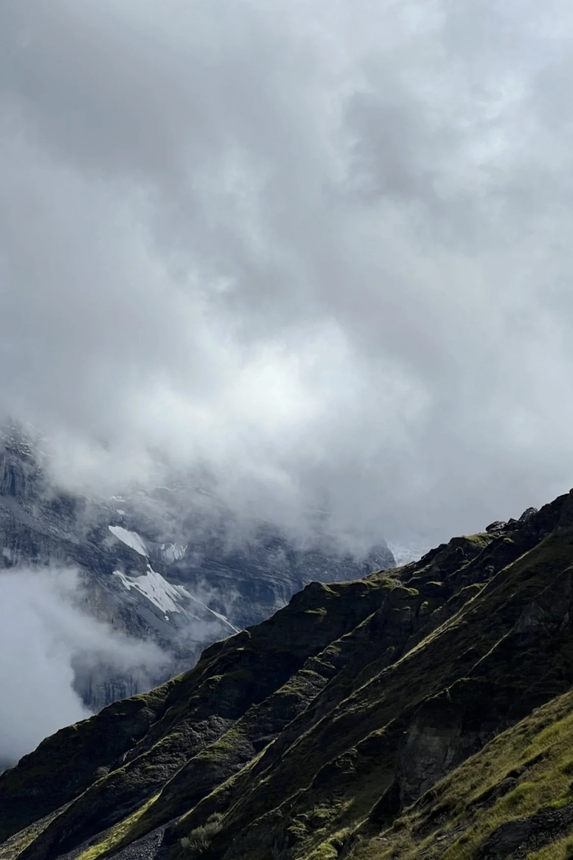Mountains with rugged cliffs, some patches of snow, and foggy, overcast sky.