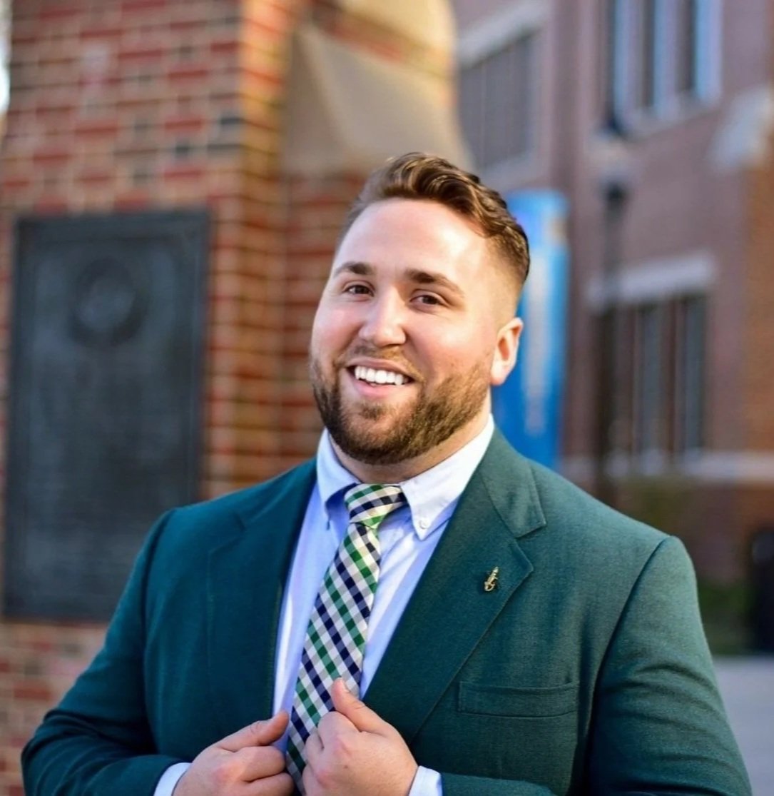 A smiling man with short brown hair, beard, wearing a dark green suit jacket, checkered tie, and white shirt standing outdoors in front of a brick building.